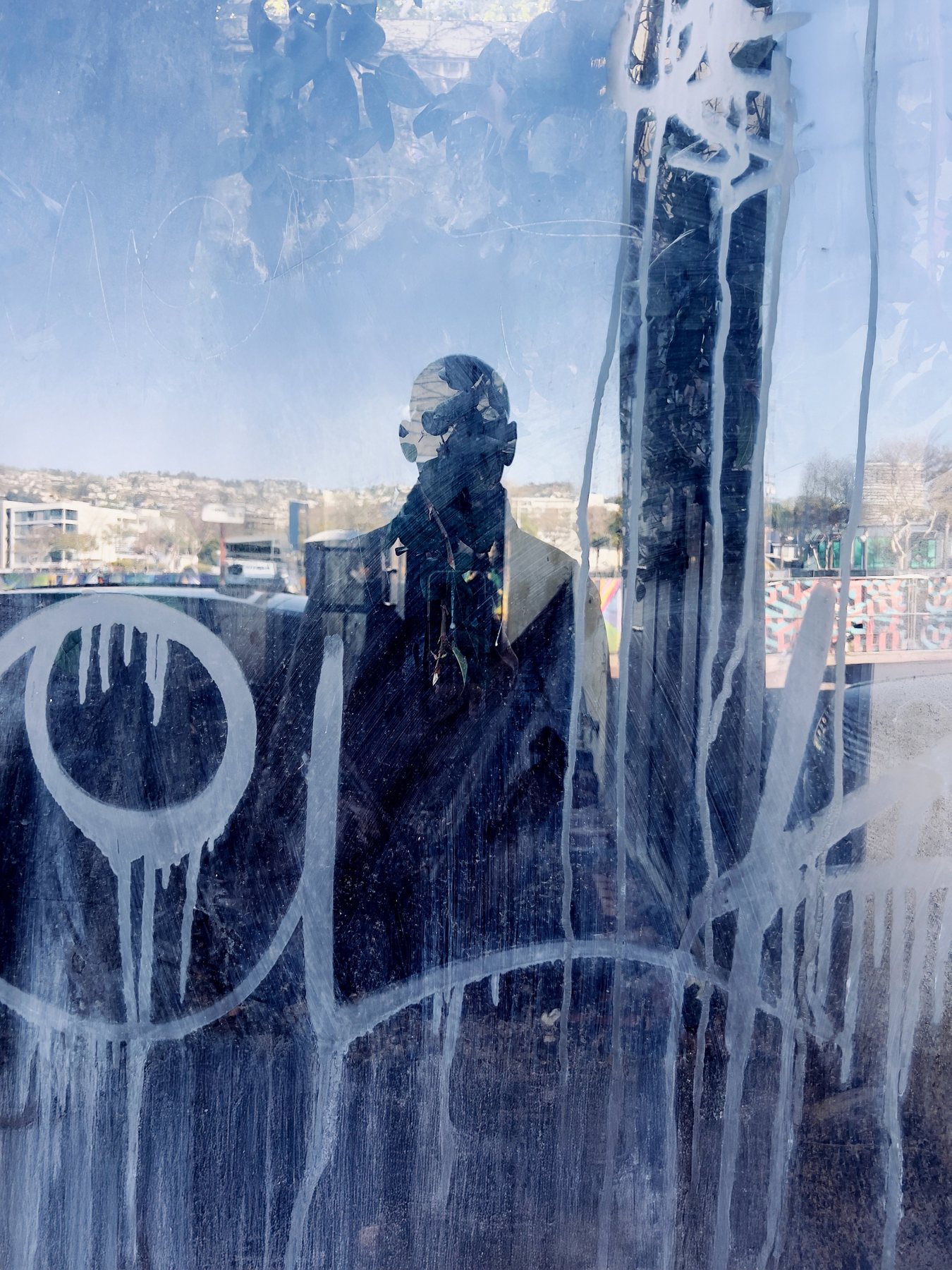 No End In Sight 072: The artist's head and shoulders reflected in blue-tinted glass covered in white paint drips and graffiti, with a hillside town behind.