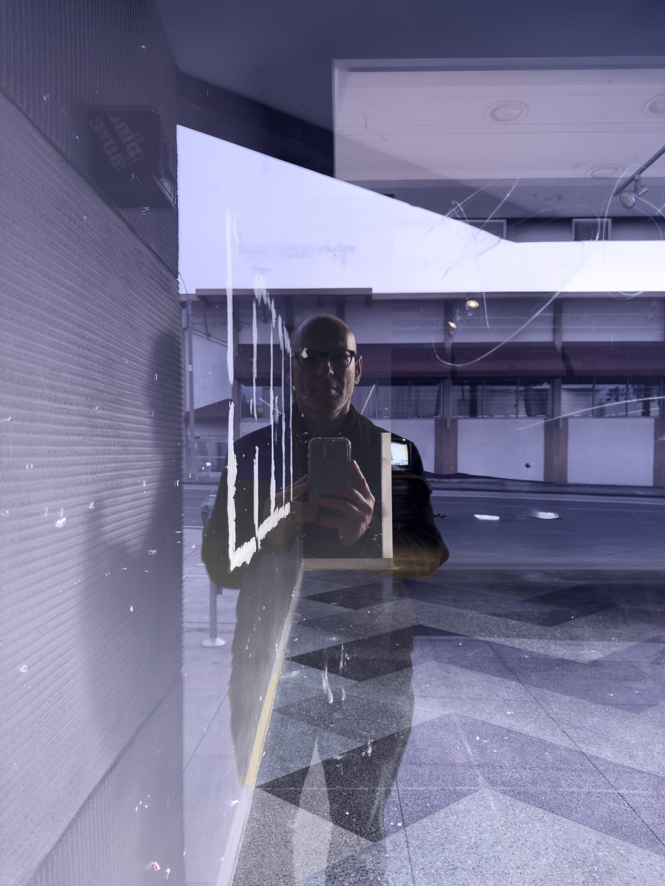 No End In Sight 132: The artist, with a phone raised, reflected in a glass corner, with a wall at left and a geometric tiled pavement below.