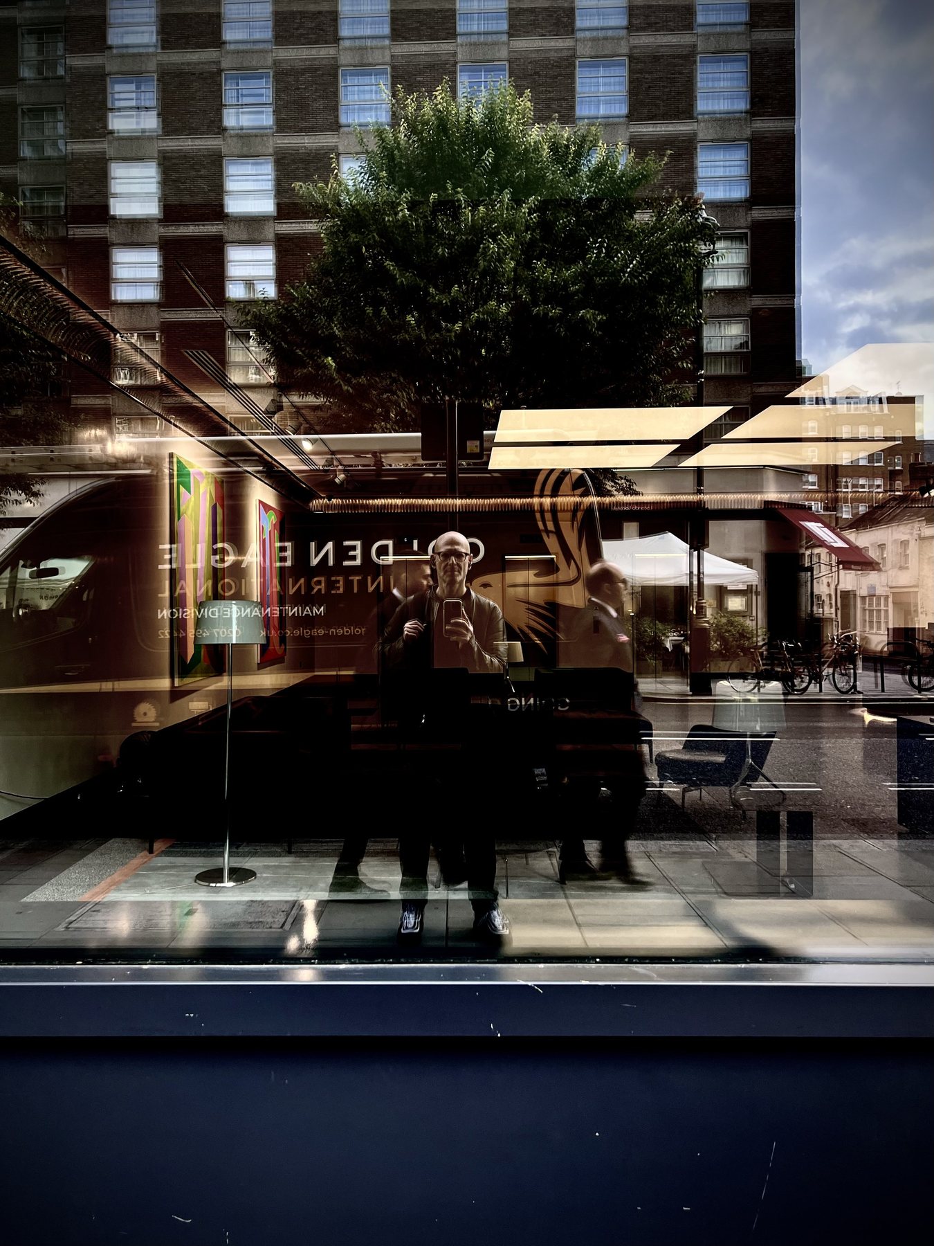 No End In Sight 021: The artist and a pedestrian reflected in a shop window with Golden Eagle signage, a tree and a tall brick building behind.