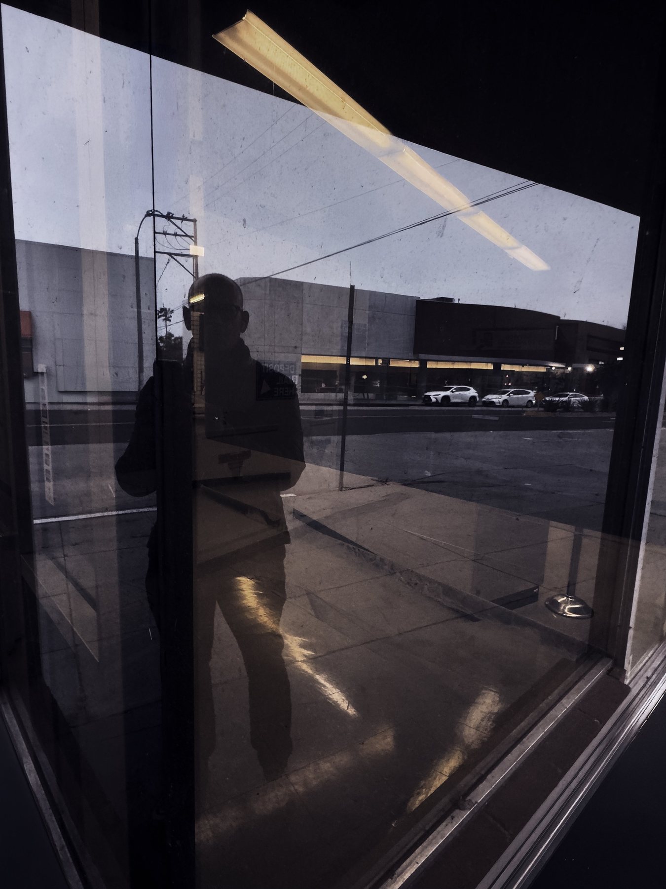 No End In Sight 127: The artist's dark silhouette reflected in a large window at dusk, with a fluorescent tube overhead and a street beyond.