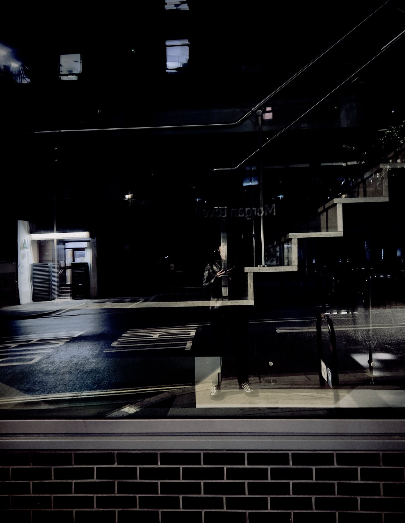 No End In Sight 019: Dark night window reflecting an angular staircase and an empty road, the artist a small figure with a phone beside a brick base.