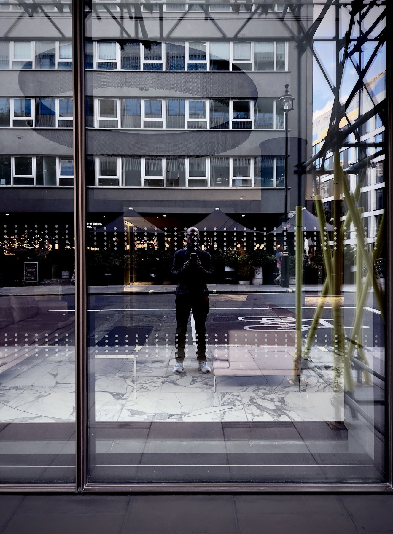 No End In Sight 046: The artist with a phone reflected in a dotted-glass window facing a grey office block and scaffolding, with a marble floor below.