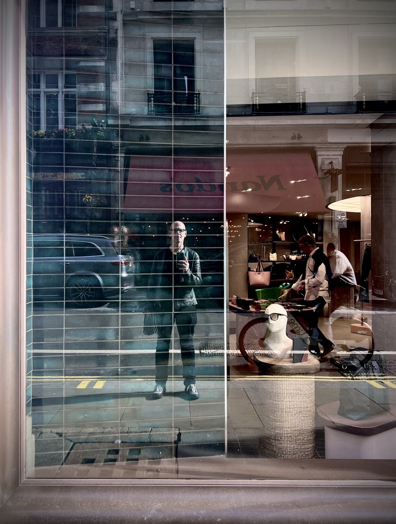 No End In Sight 029: The artist with a phone reflected in a window with a Nardos sign, blinds, mannequin, shoppers and a parked car.
