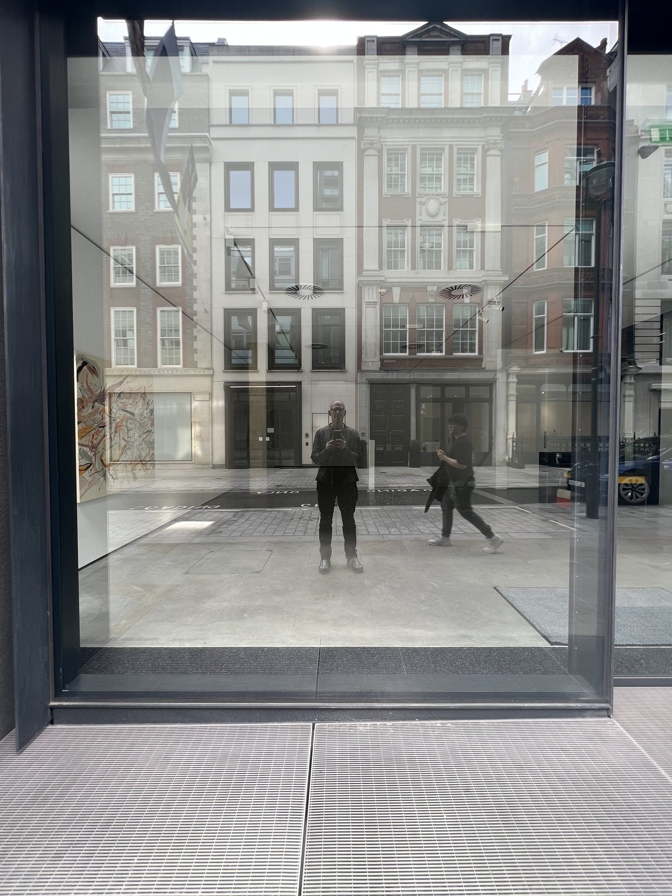 No End In Sight 027: The artist with a phone reflected in a gallery-style glass window, with a pedestrian crossing a pale street lined with buildings.
