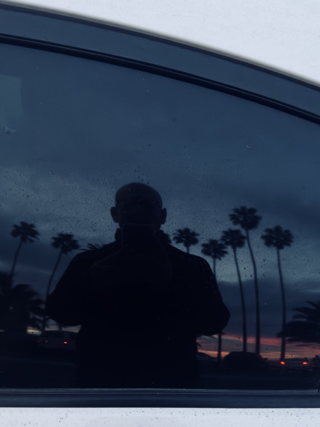 No End In Sight 096: The artist's silhouette in a dark car window at dusk, with palm trees and an orange sunset reflected and raindrops on the glass.