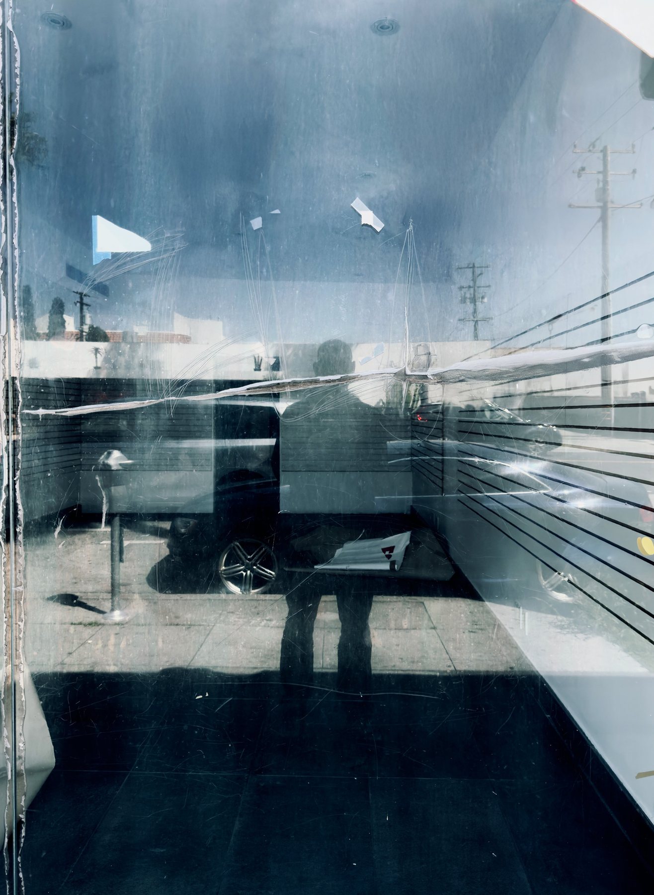 No End In Sight 131: The artist's reflection in a scratched, cracked window with a blue sky ceiling, utility poles, a car and a striped wall.