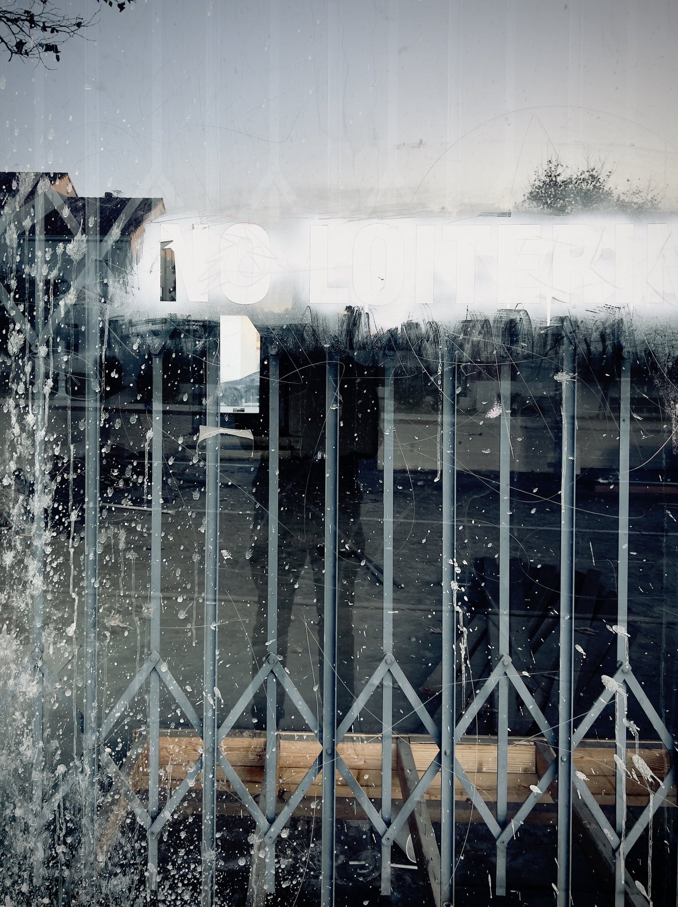 No End In Sight 062: White paint and splashes across a window with a folding security grille, with a dim silhouette reflected behind it.