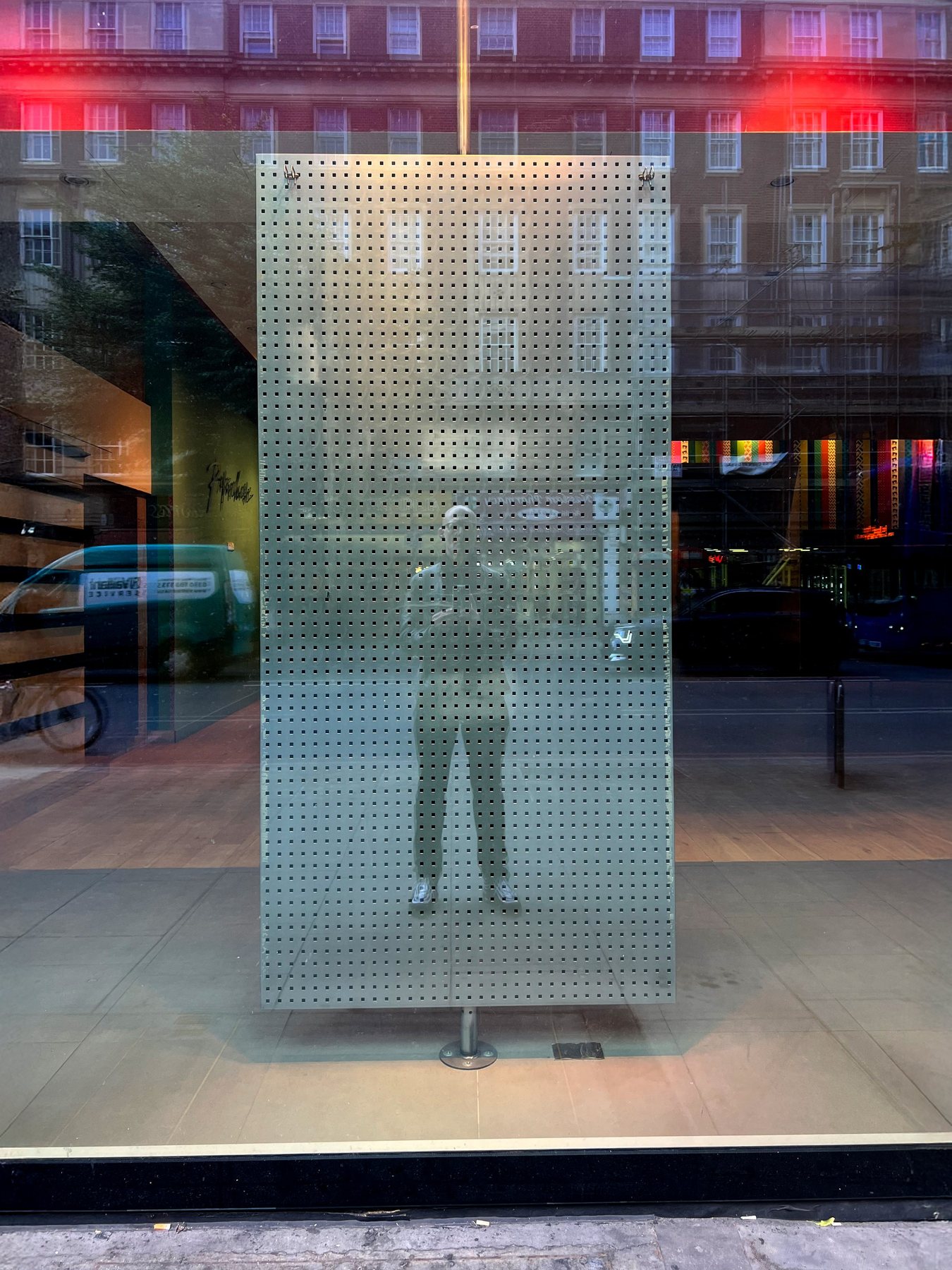 No End In Sight 004: The artist reflected in a pegboard-like perforated glass panel in a shop window, with a street, red light and a green van behind.
