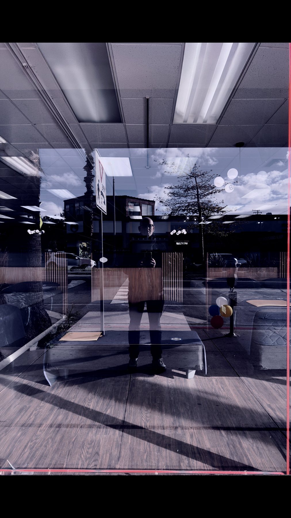 No End In Sight 123: The artist's tiny reflection deep in a layered shop window, with fluorescent ceiling lights, balloons and sunlit clouds.