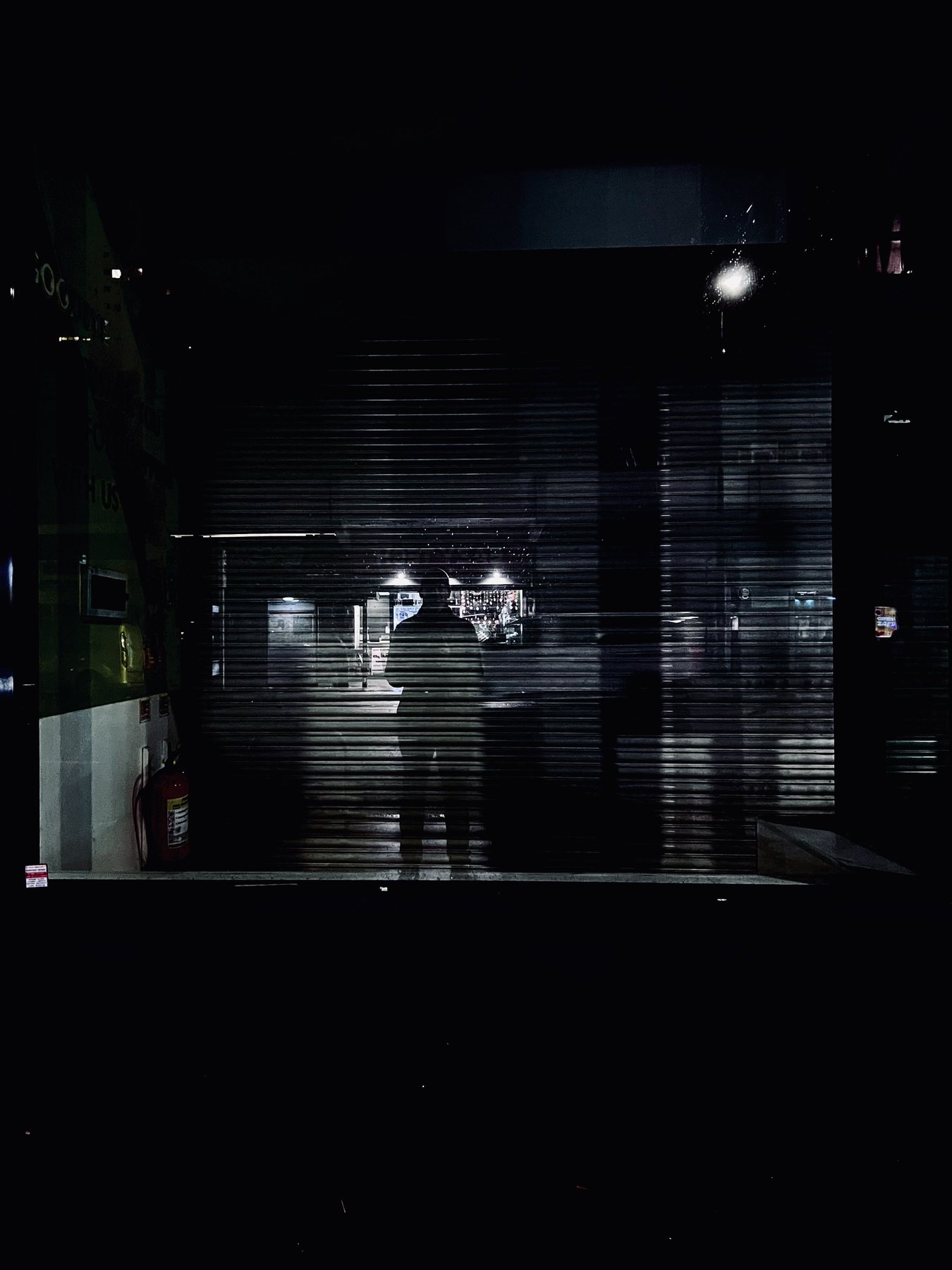 No End In Sight 015: The artist's shadowy figure behind a closed roller shutter in a dark space with lights beyond and a fire extinguisher at left.