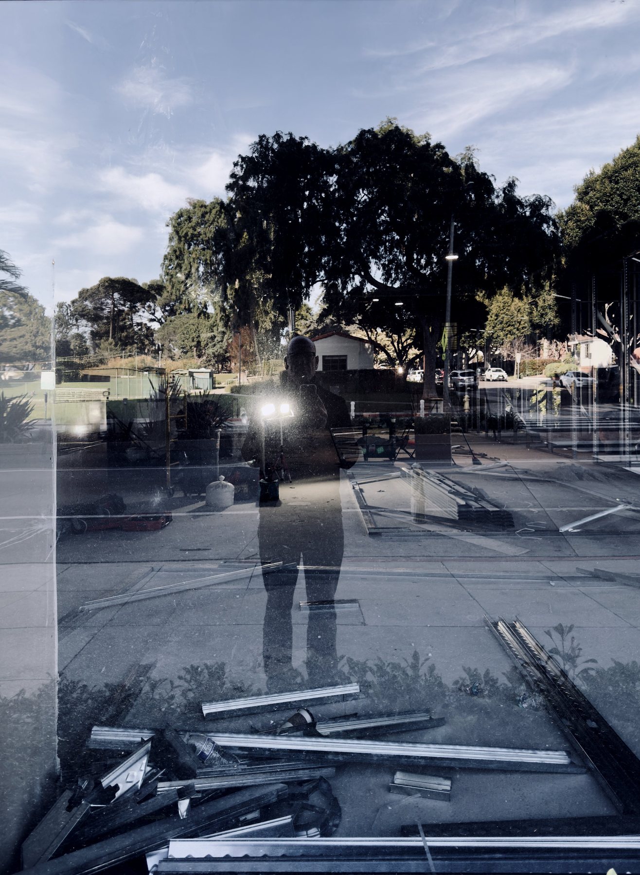 No End In Sight 031: The artist's dark silhouette in a window overlooking a tree-lined street under blue sky, with scattered metal frames and debris in the foreground.