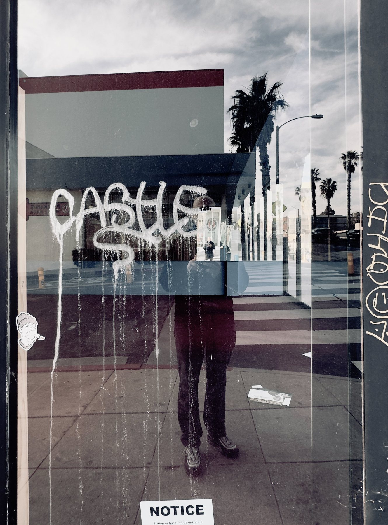 No End In Sight 129: The artist reflected beneath white drip-style graffiti reading CASTLE on glass, with palm trees, a street corner and a NOTICE sign.