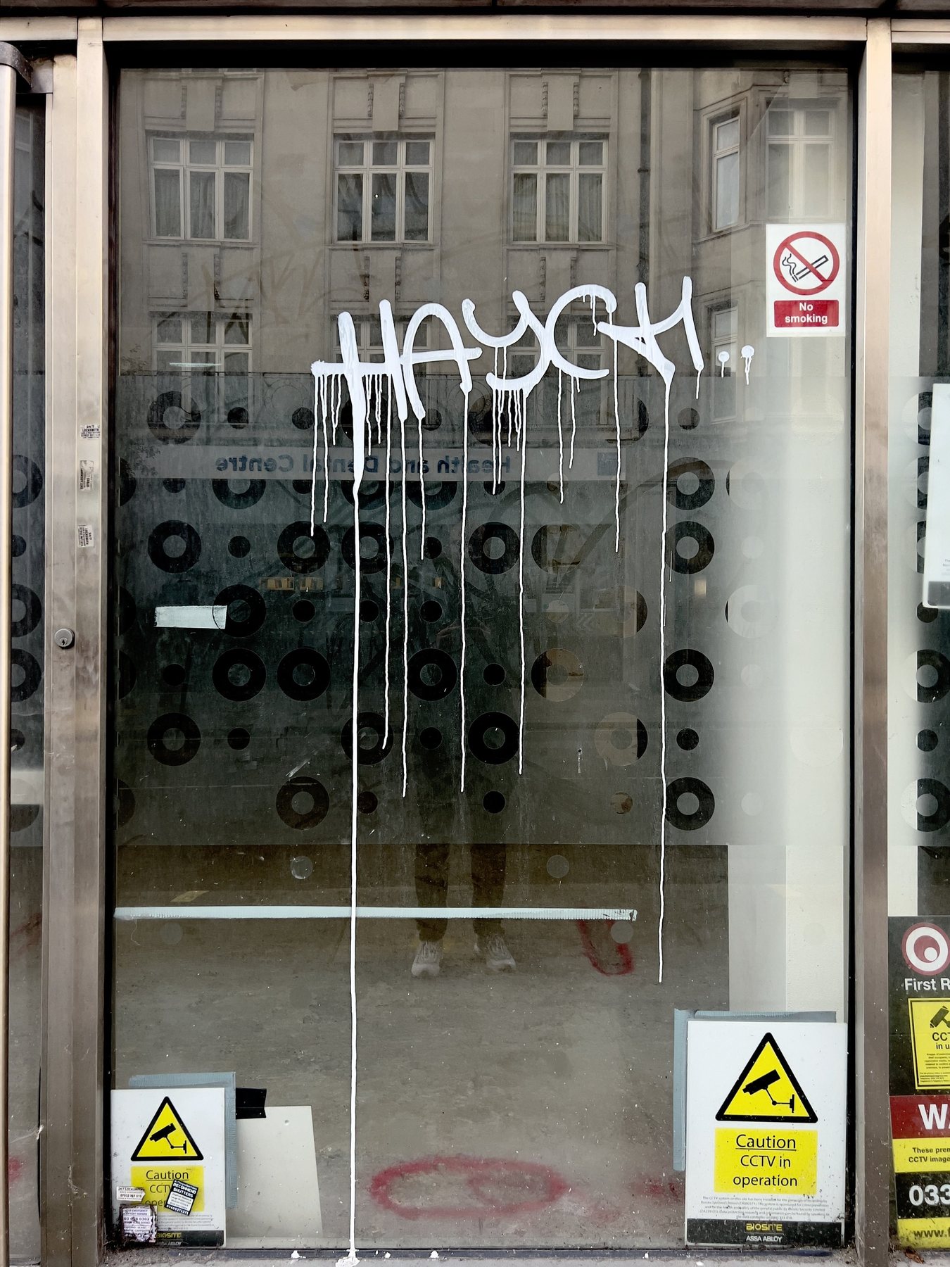 No End In Sight 047: A glass door with white graffiti reading HAYGH dripping over a dotted pattern, with CCTV signs and the artist's lower legs.