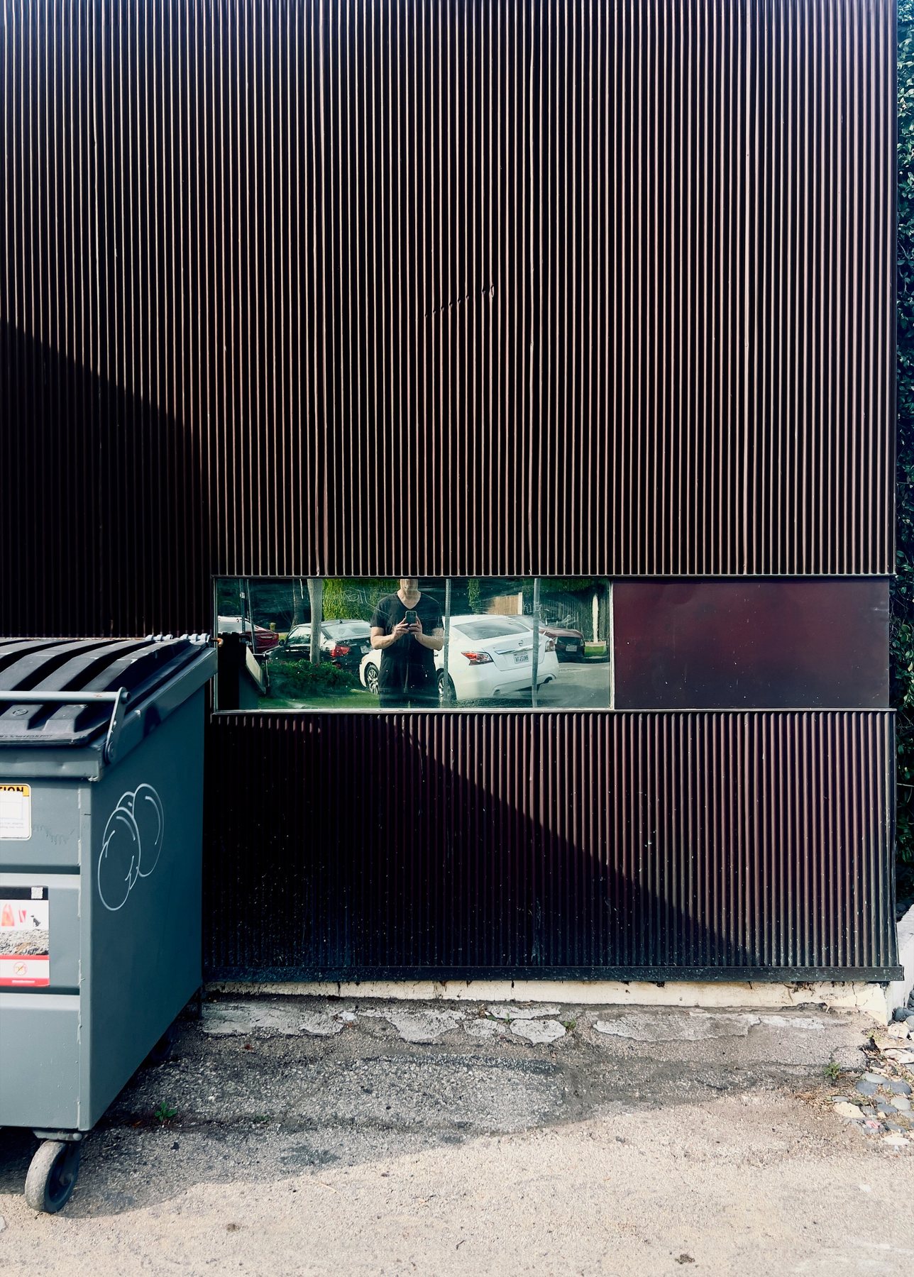 No End In Sight 066: The artist photographing himself in a narrow glass strip set in a dark corrugated wall, beside a green dumpster.