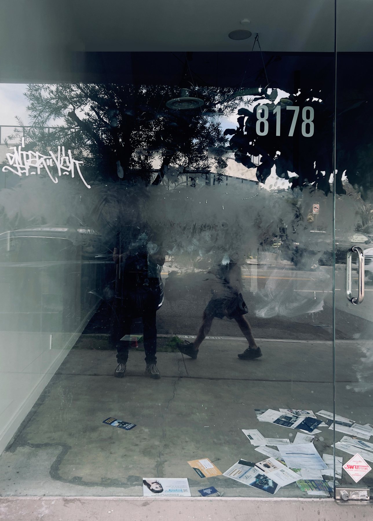 No End In Sight 070: A glass door with number 8178 and the artist beside another passerby, reflecting a tree, with flyers scattered on the floor.