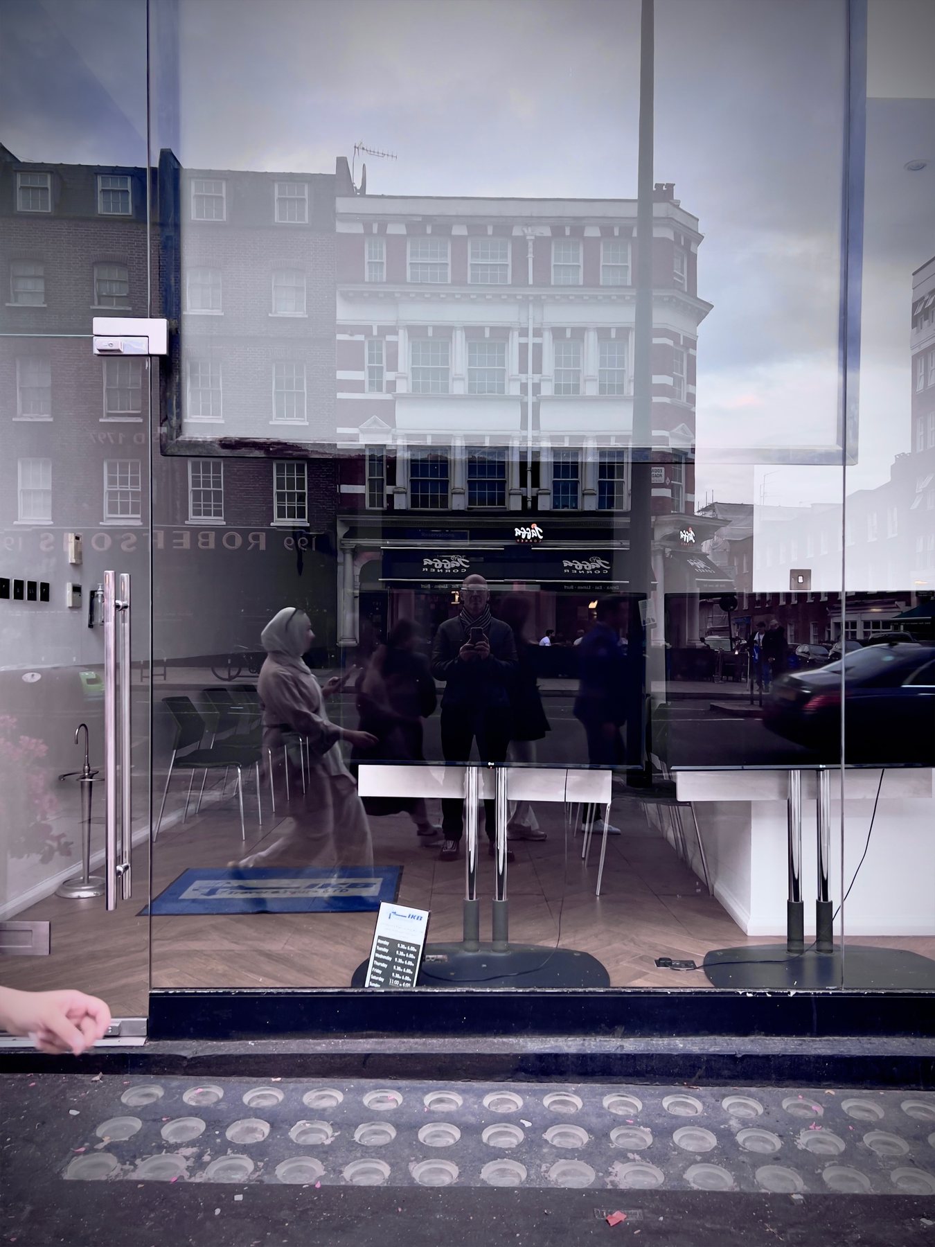 No End In Sight 032: The artist's reflection in a glass shopfront, layered over brick buildings, a passing woman in a headscarf and tactile paving.