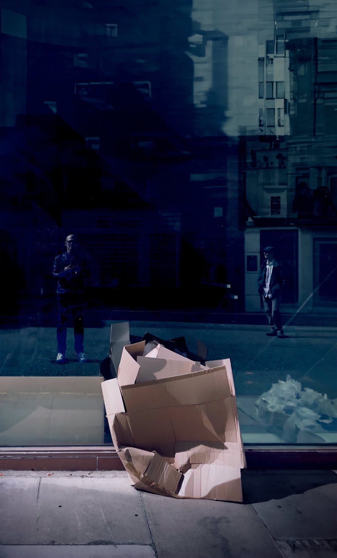 No End In Sight 053: Flattened cardboard on a pavement before a dark window in which two figures and city buildings are reflected.