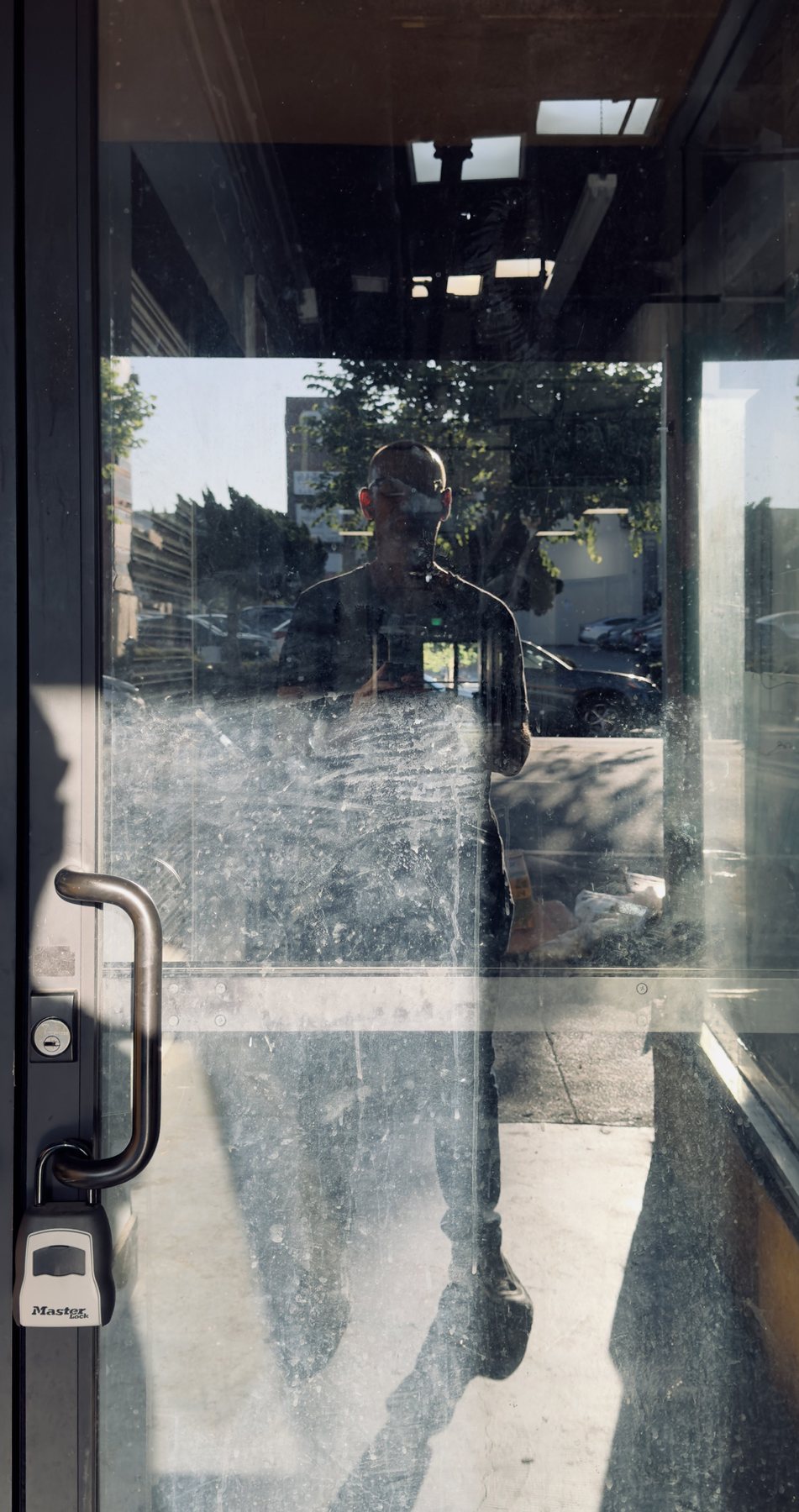 No End In Sight 081: The artist with sunglasses reflected in a scratched glass door with a metal handle and padlock box, with trees and a car behind.