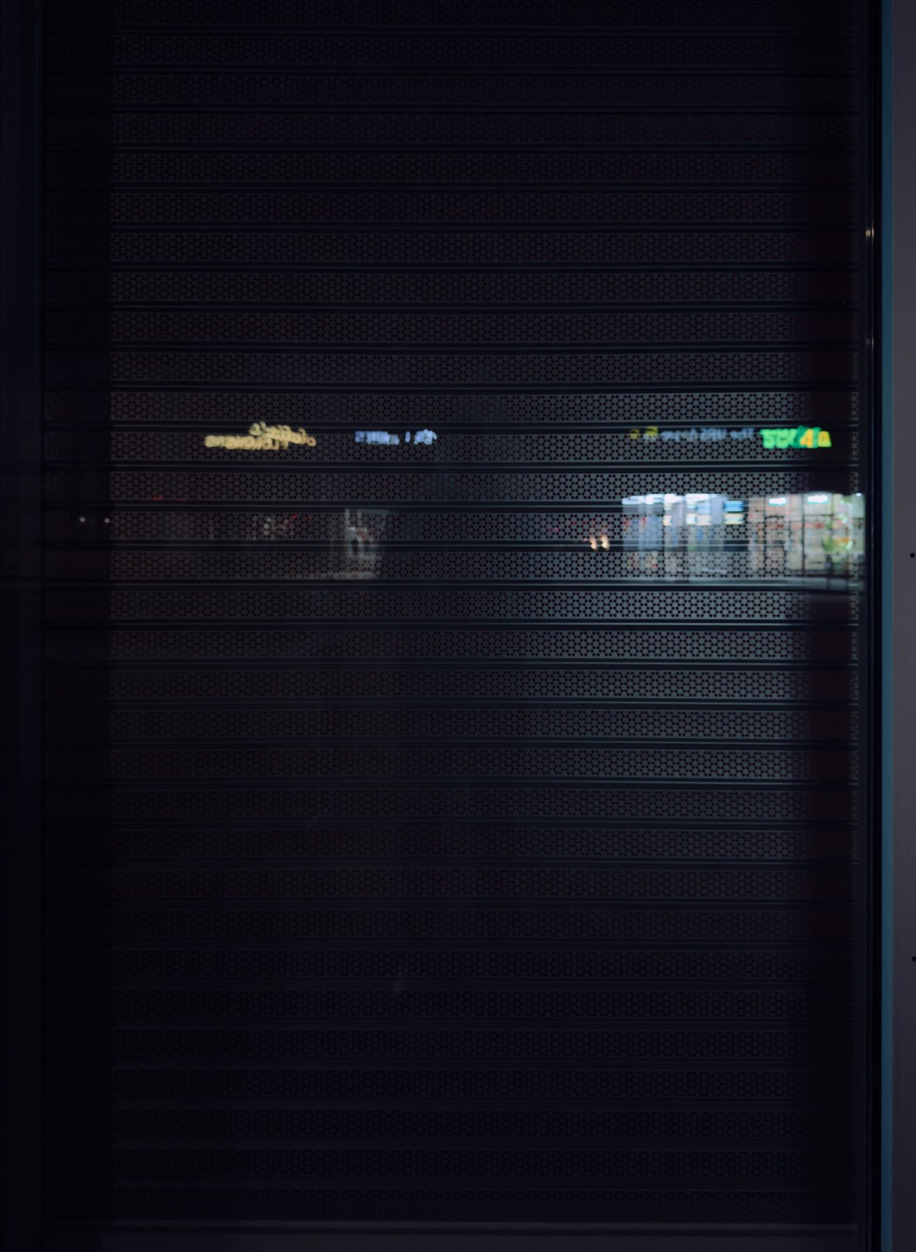 No End In Sight 103: A dark roller shutter with faint coloured shop-sign reflections along a horizontal band, no figure visible.