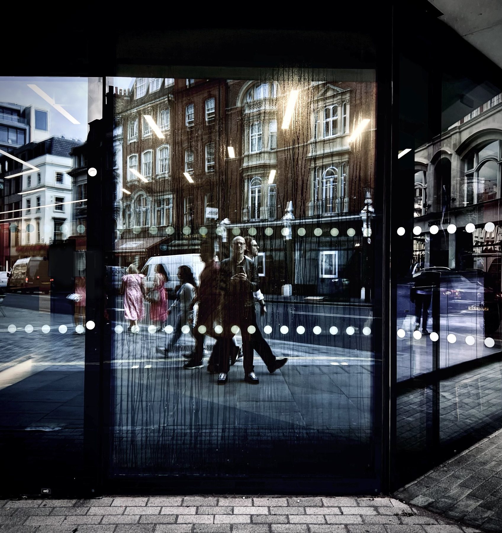 No End In Sight 024: The artist and several pedestrians, including two in pink, reflected in a window with dotted glass and brick buildings.