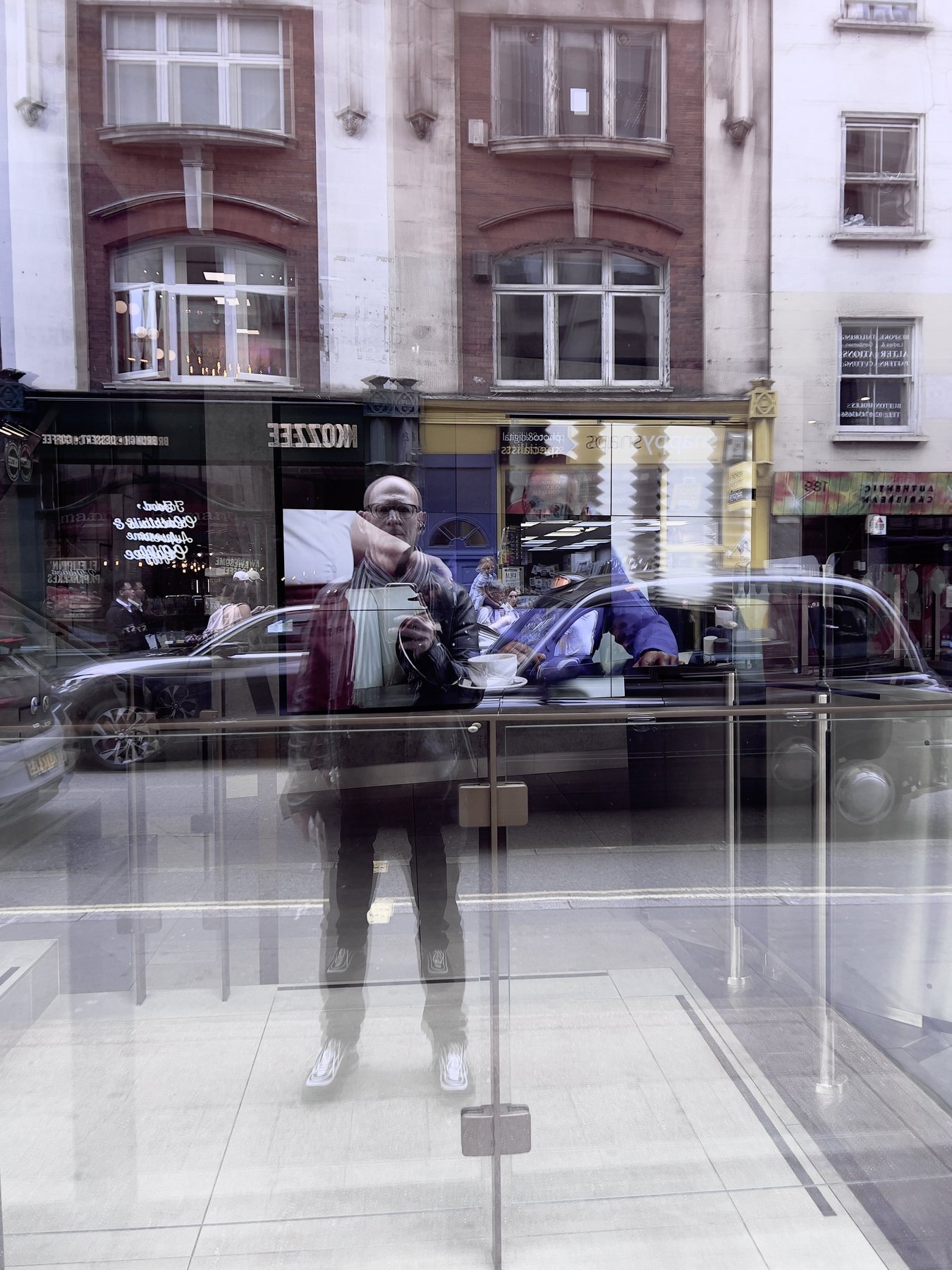 No End In Sight 050: The artist taking a photo, reflected in a glass storefront and balustrade on a London street, with brick buildings and a car.