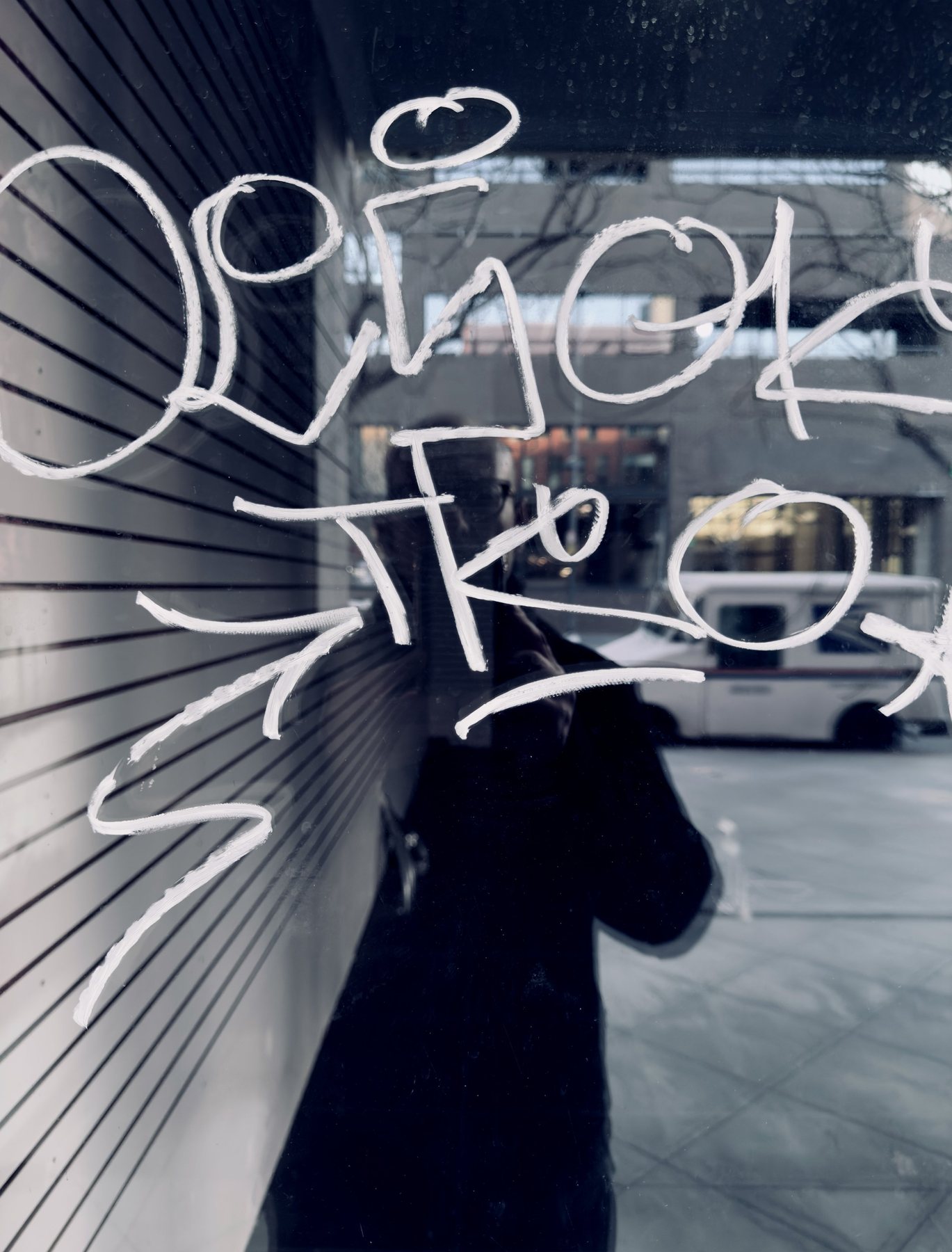 No End In Sight 116: Close view of white marker graffiti on a window, with the artist and a white delivery van reflected faintly behind.