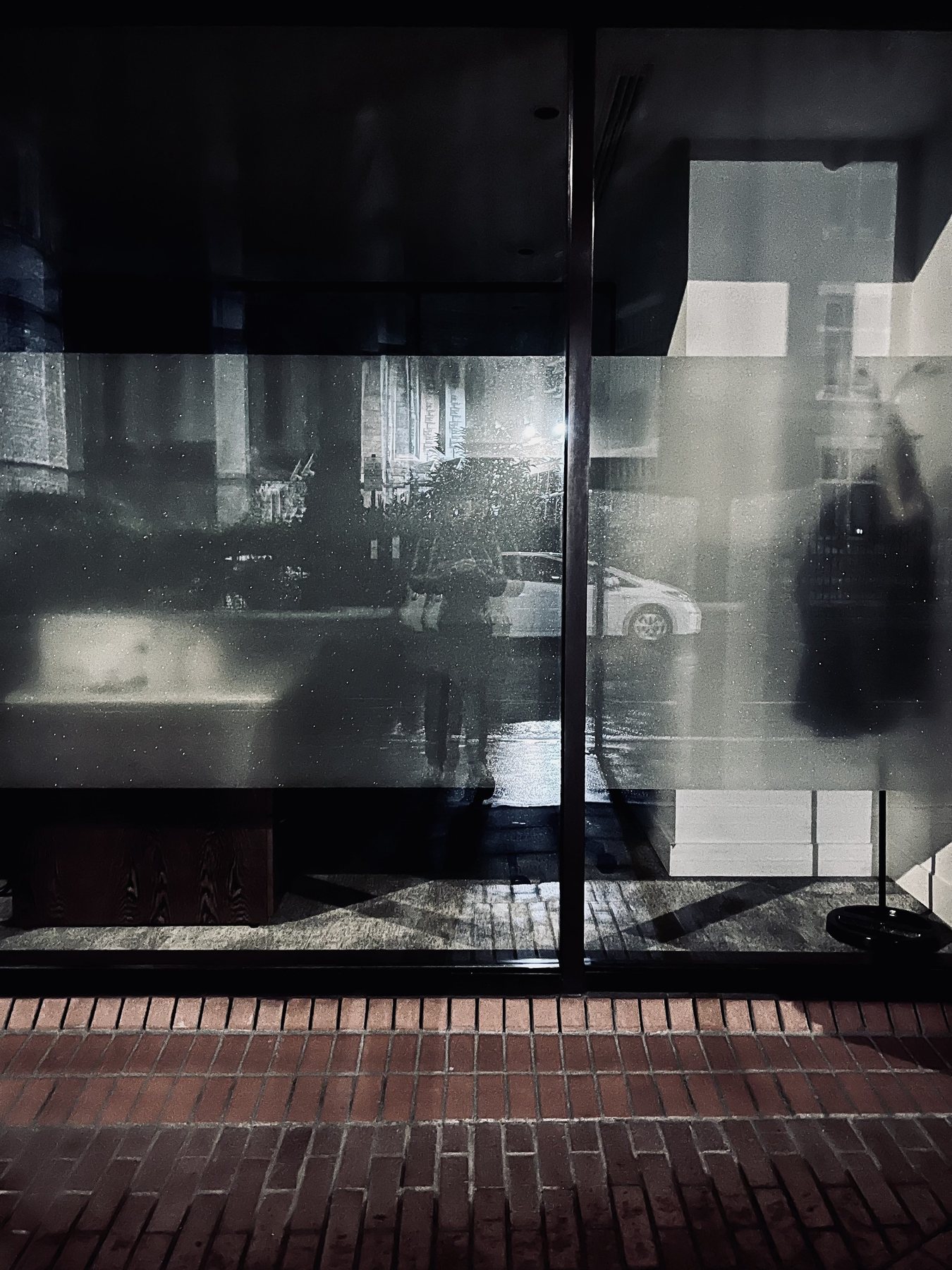 No End In Sight 016: A rainy night window with frosted glass band reflecting a white taxi, trees, and the artist, above a red brick pavement.