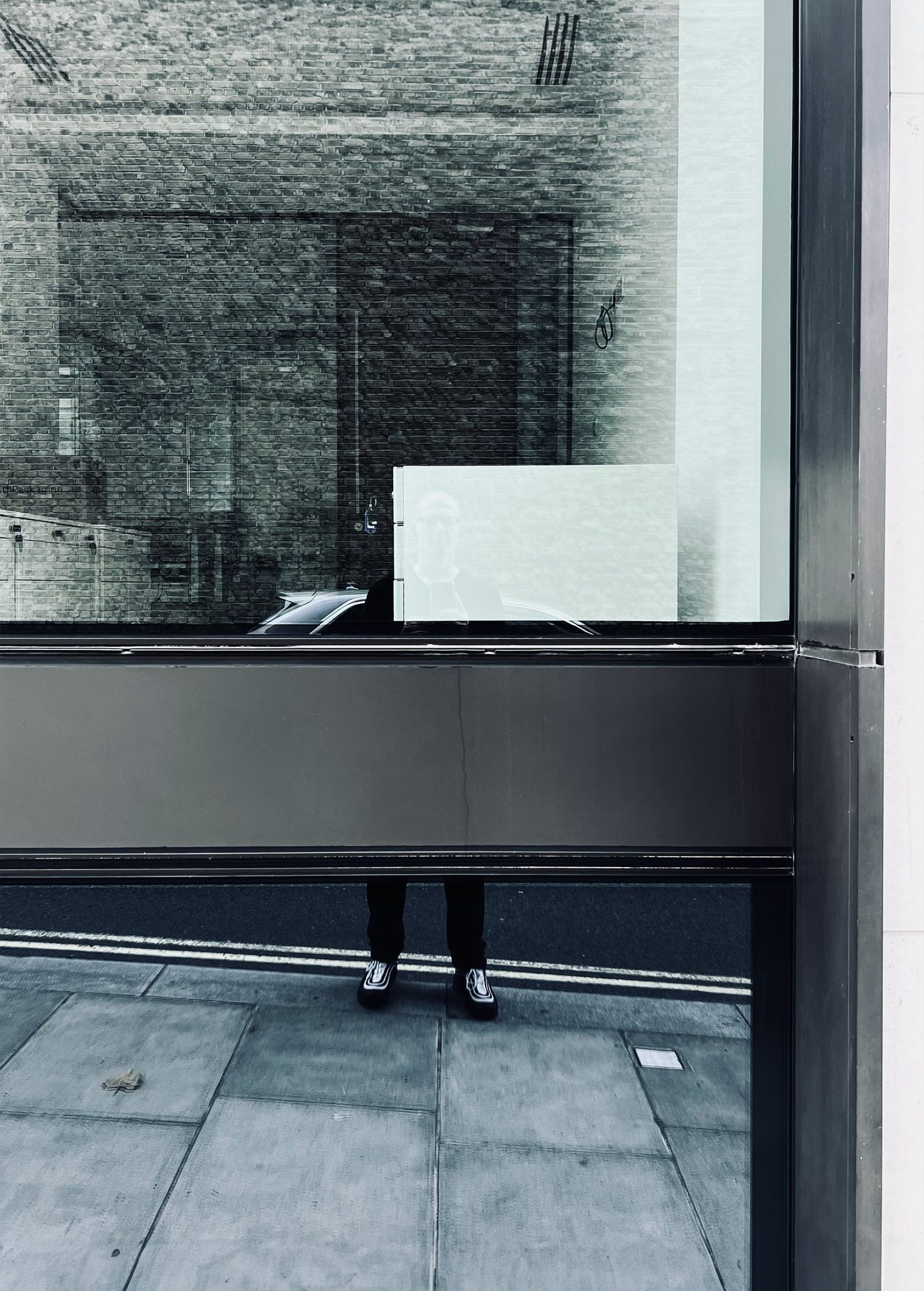 No End In Sight 034: The artist's legs and shoes visible below a frosted panel, with a reflected brick facade and a blank white sign in the window.