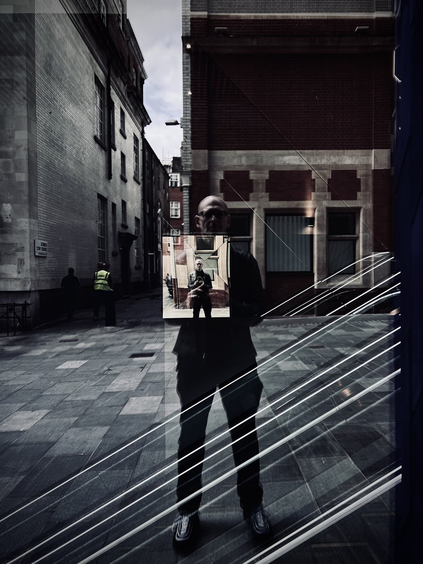 No End In Sight 008: The artist reflected in a window holding a small mirror that shows another reflection of the artist, in a narrow brick alley.