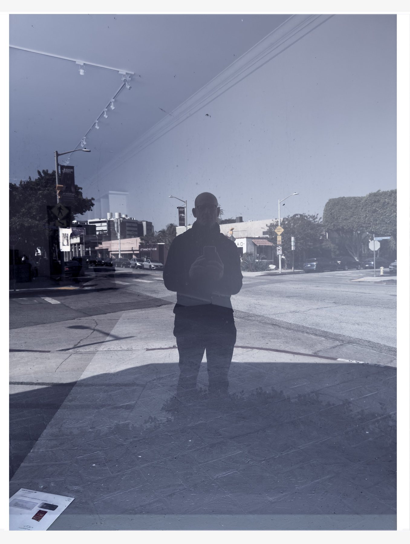 No End In Sight 143: The artist's silhouette reflected in a blue-grey window above a wide street, with ceiling track lights reflected at top left.
