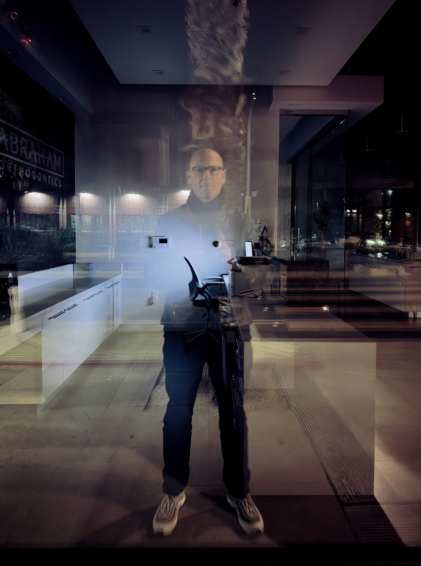 No End In Sight 104: The bespectacled artist reflected in a night-time window, with a palm trunk, a clinic counter and white trainers visible.