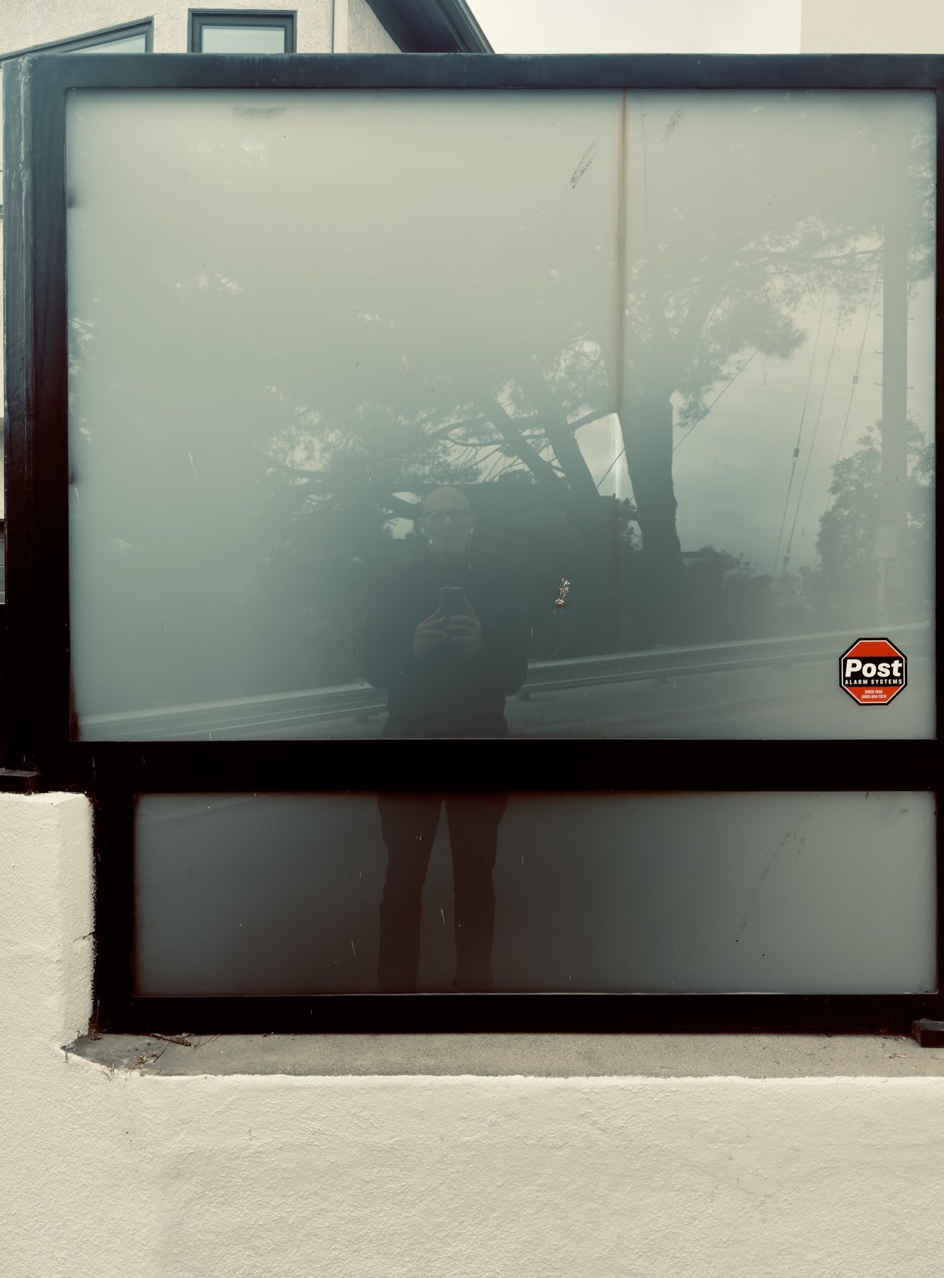 No End In Sight 079: The artist with a phone reflected in a frosted window with grey sheen, trees and a road behind, and a security alarm sticker on the glass.