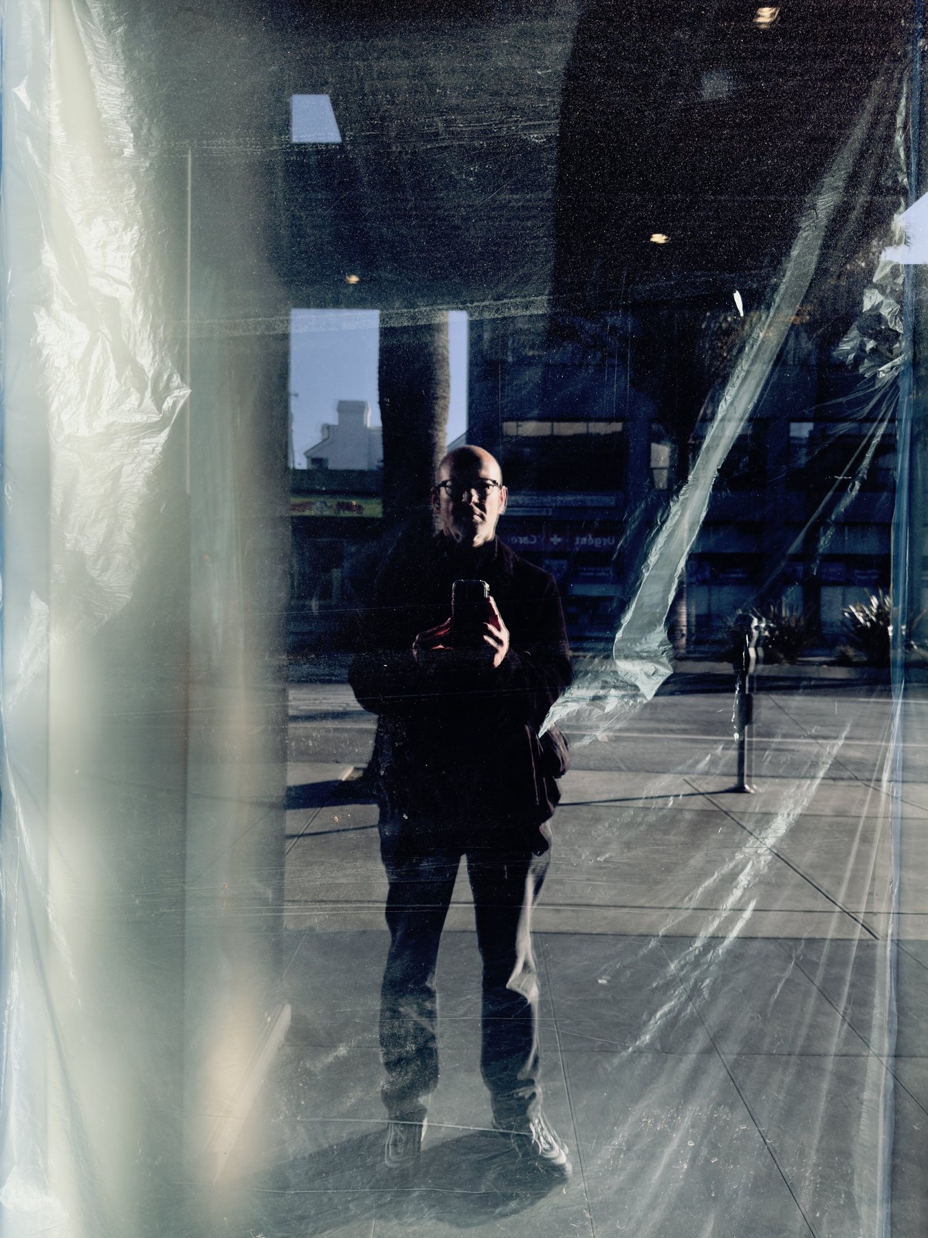 No End In Sight 112: The artist with a phone reflected in a window draped with clear plastic sheeting, with low sun on a paved street and a parking meter.