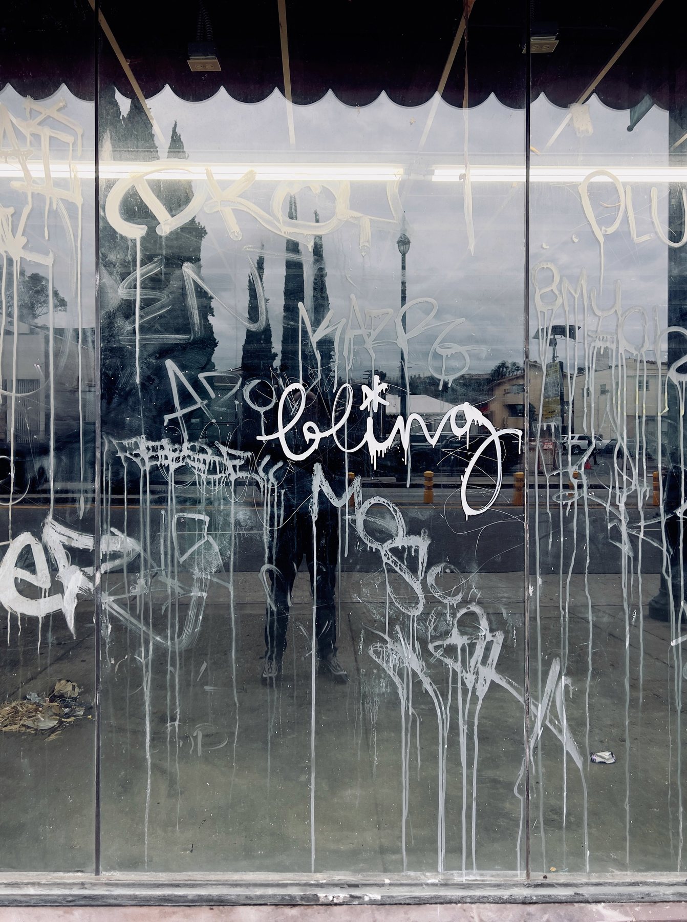 No End In Sight 071: Bling: the artist's reflection in a shop window scrawled with white graffiti tags and drips, with the word "bling" at centre.