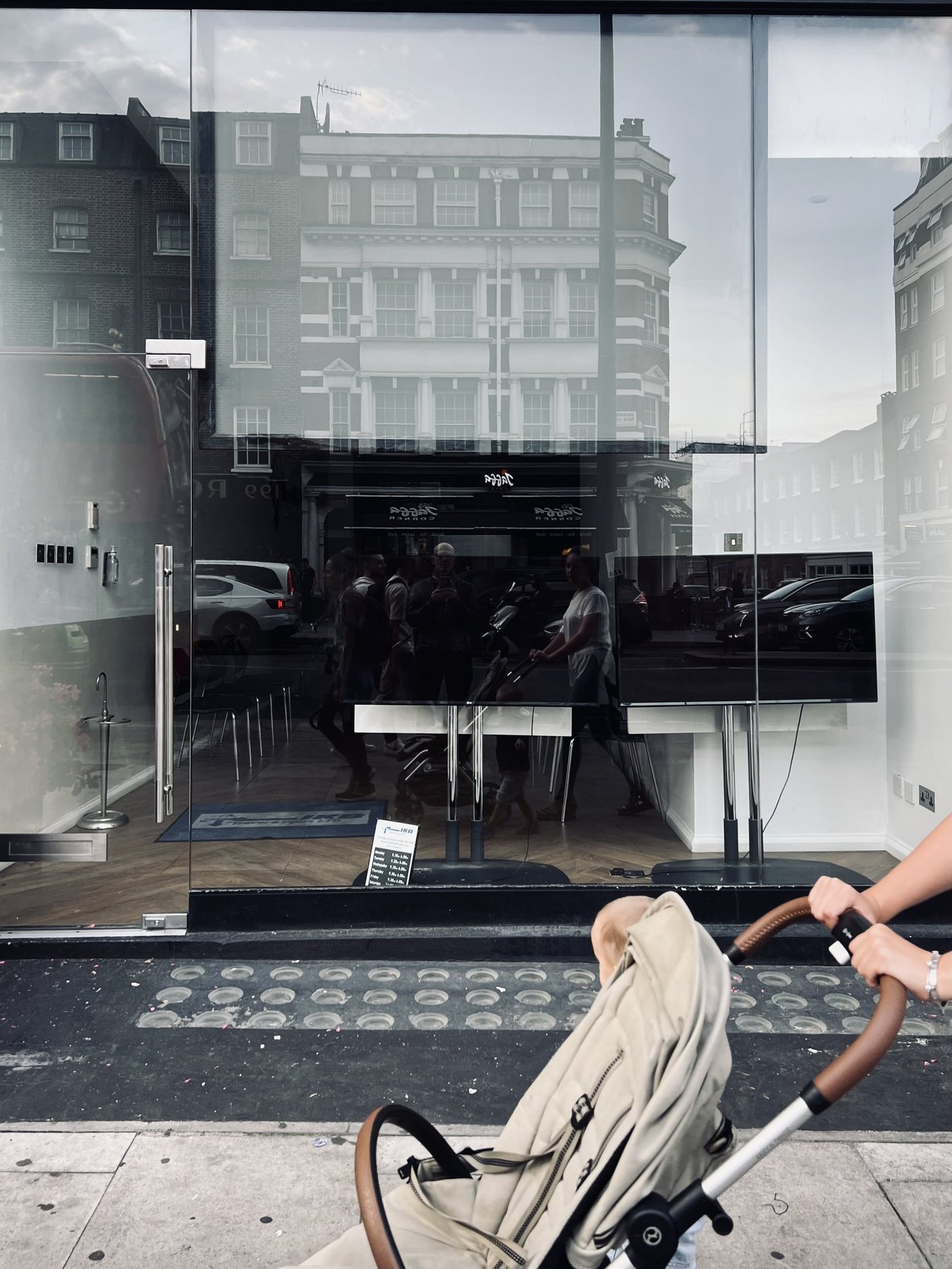 No End In Sight 030: A shop window reflecting terraced buildings and a TV screen with a pram's beige canopy and a parent's hand in the foreground.