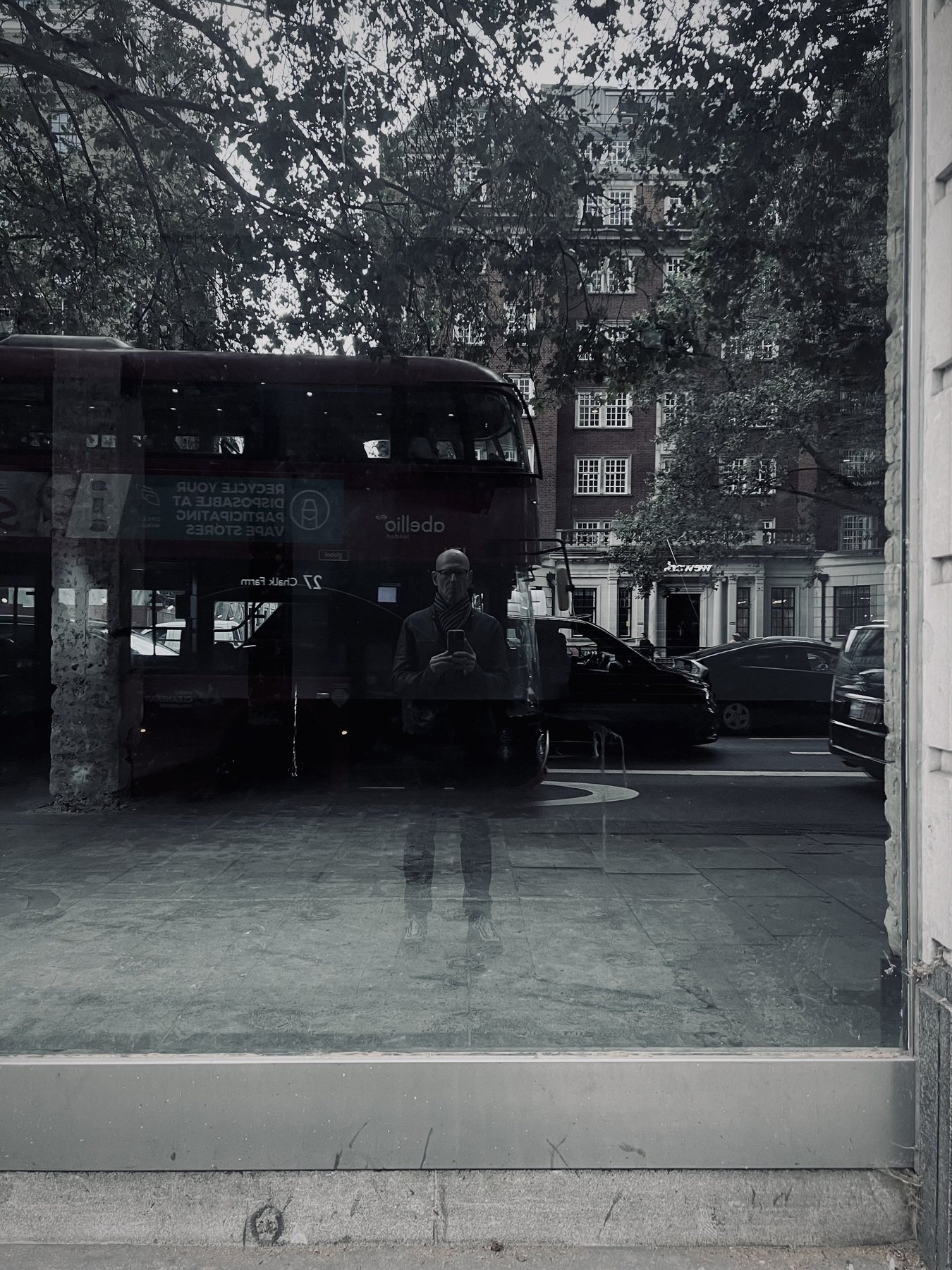 No End In Sight 040: The artist reflected in monochrome window glass with a red double-decker bus, trees and a dark car behind.