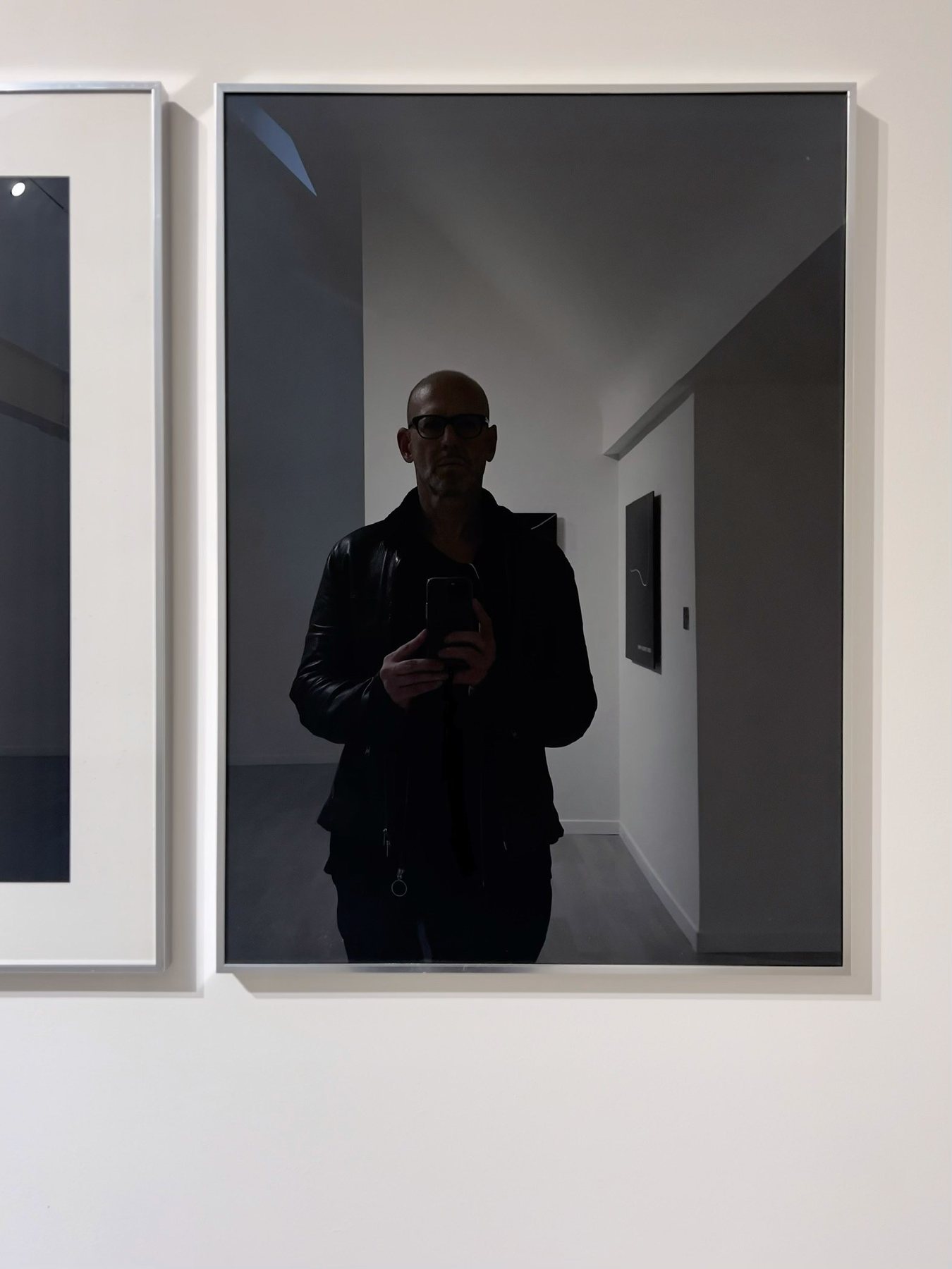 No End In Sight 054: Close view of the framed mirror in a gallery, showing the artist in glasses and a leather jacket holding a phone, with a corridor behind.