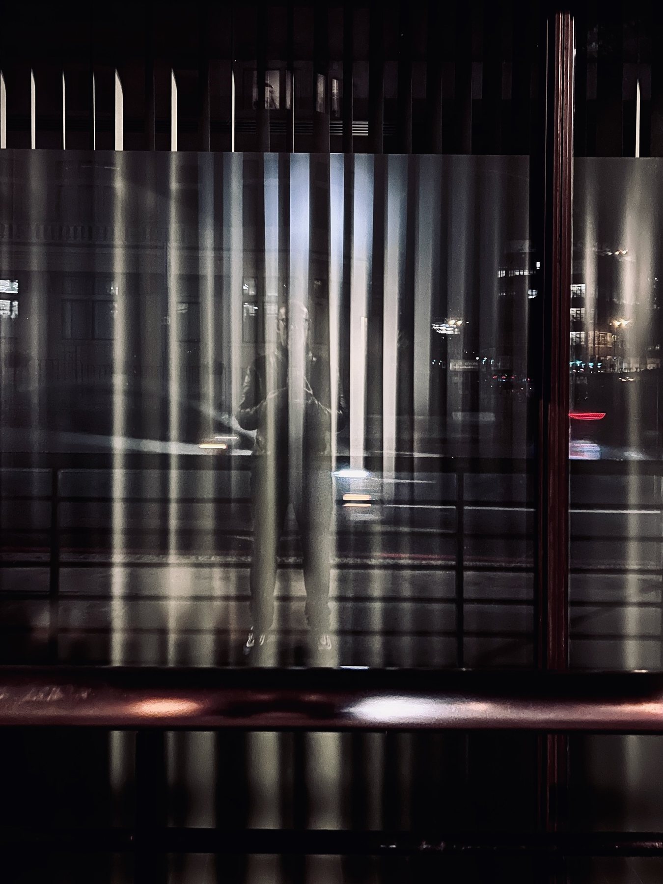 No End In Sight 036: A night street reflected in vertically ribbed glass, with the artist's blurred standing figure seen through bright streaks of light.