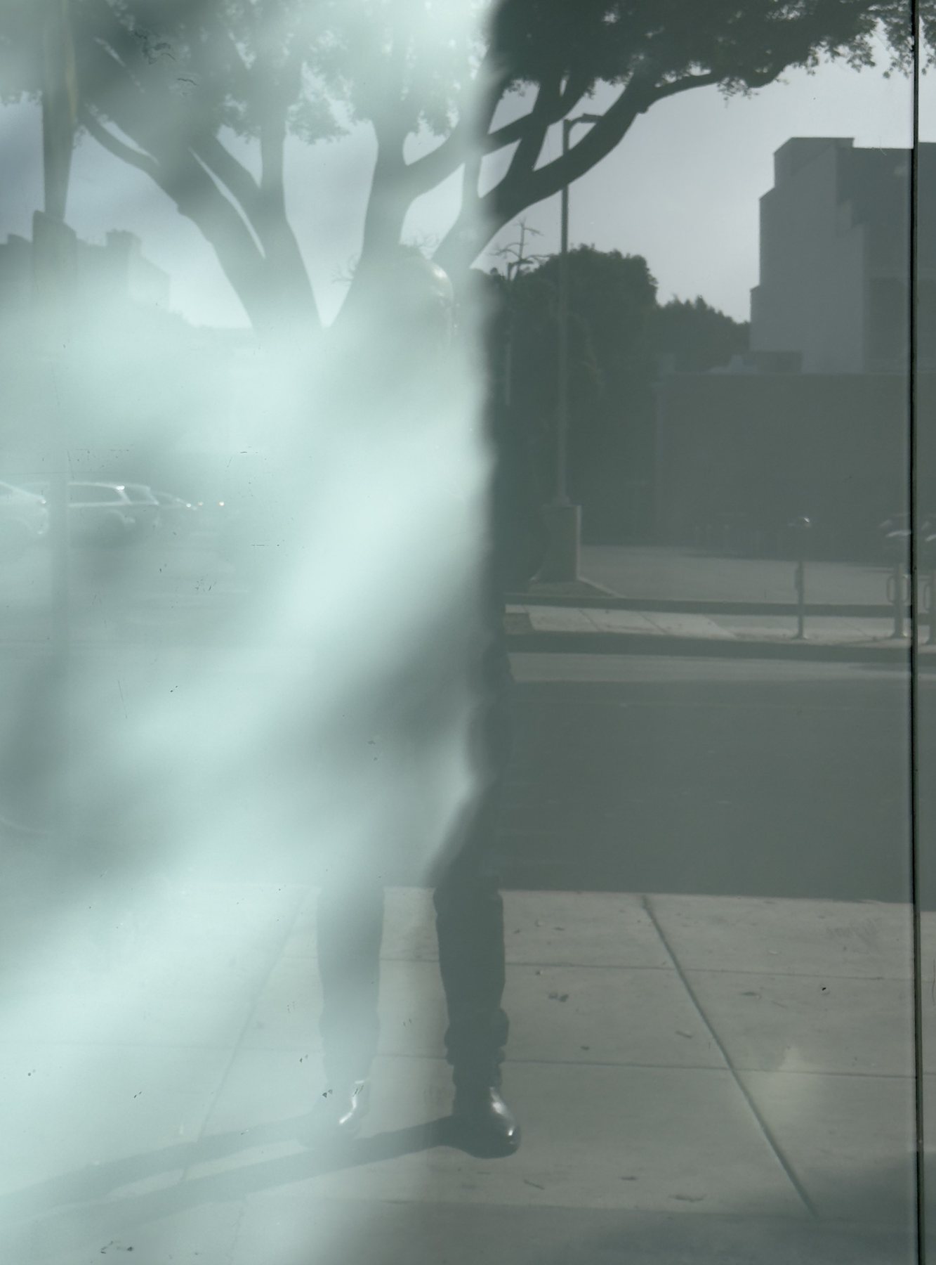 No End In Sight 099: A figure's reflection fading behind a bright milky smear on glass, with trees, pavement and a distant building.