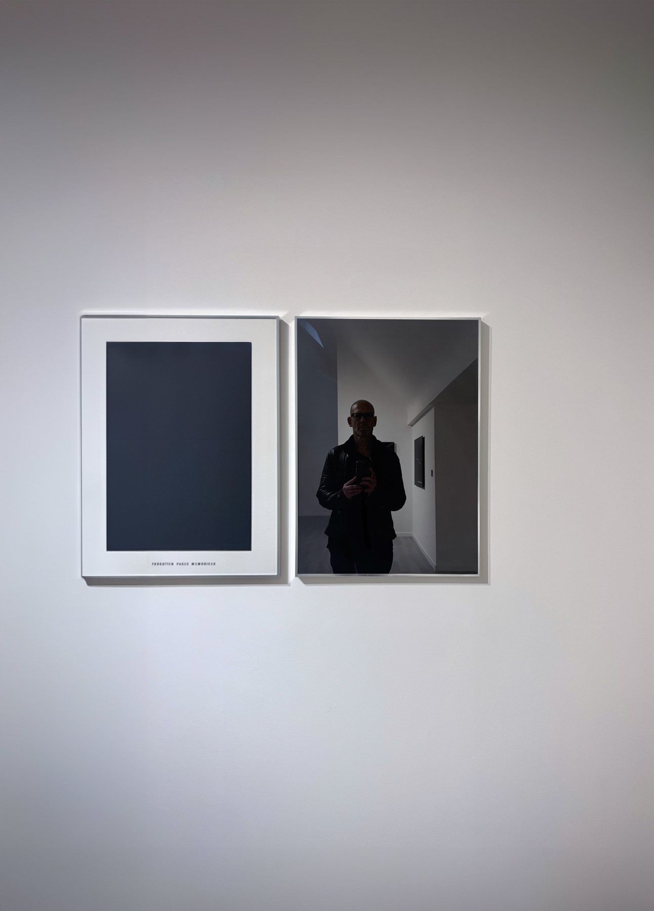 No End In Sight 055: Two framed pieces on a white wall, a dark blue print and a mirror reflecting the artist with a phone in a gallery corridor.