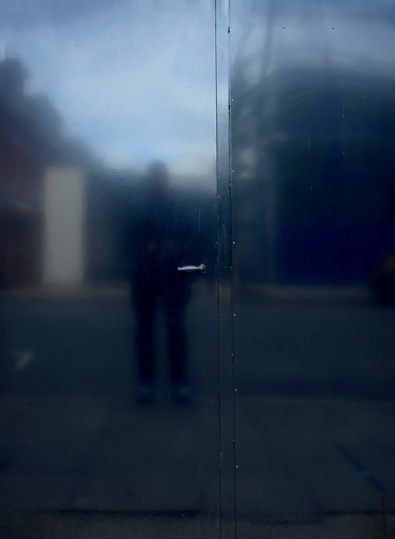 No End In Sight 060: The artist's dark silhouette reflected in a dark blue metal door with a small handle, with a hazy sky and buildings.