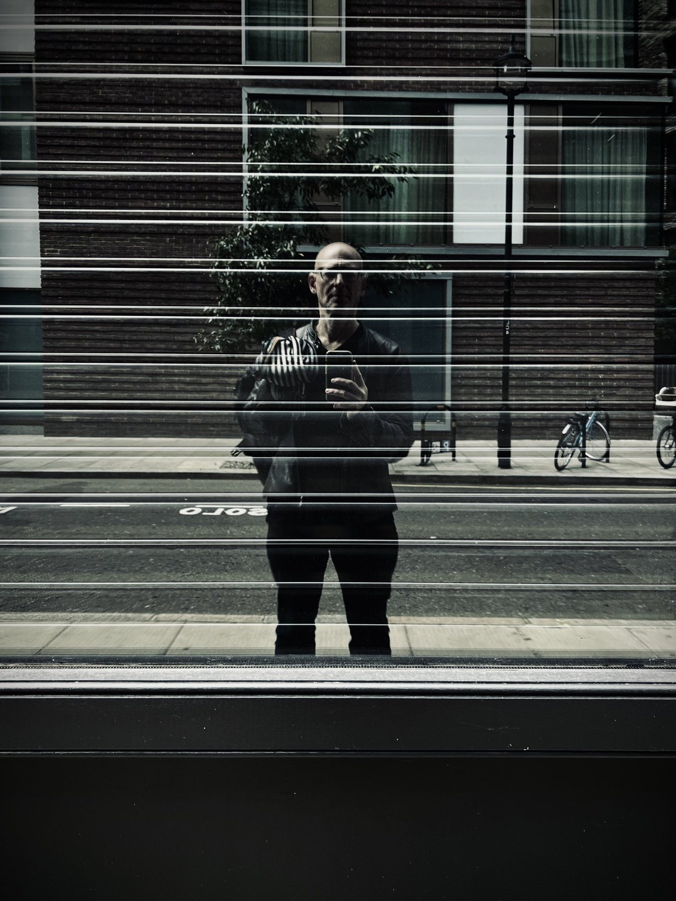 No End In Sight 039: The artist with a phone reflected through horizontal blind lines over a brick building, a street lamp and bikes.