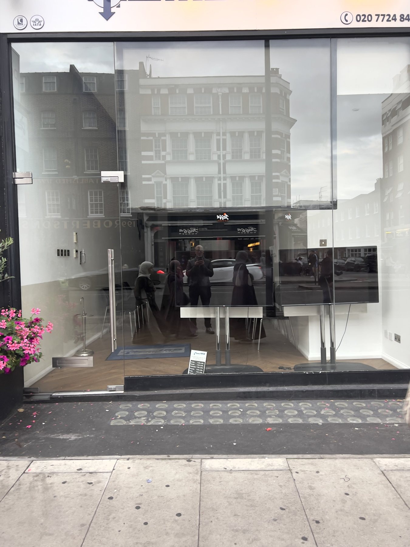 No End In Sight 033: The artist and several other reflections in a London shopfront window, with brick facades above and a pavement of paving studs below.