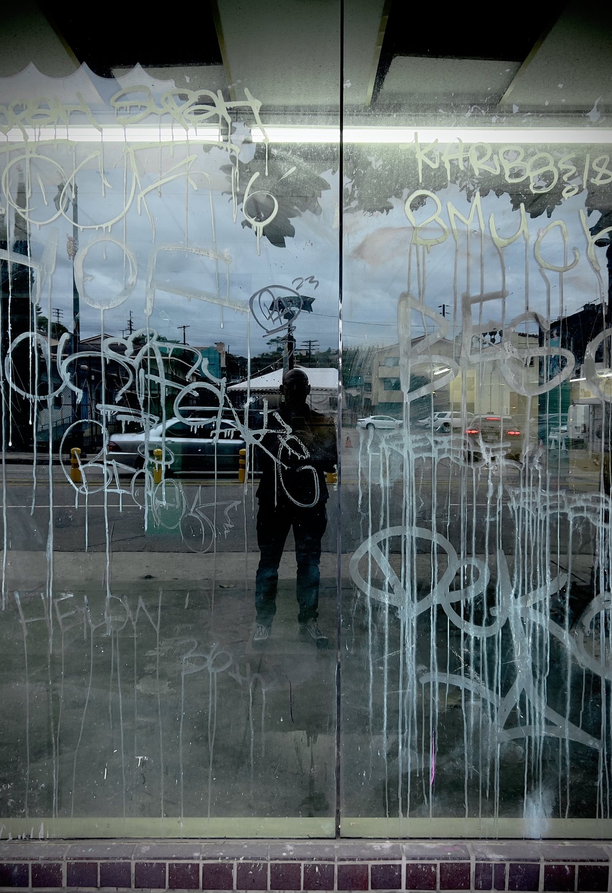 No End In Sight 057: The artist's reflection in a double glass door scrawled with white graffiti and drips, with a street and parked cars beyond.