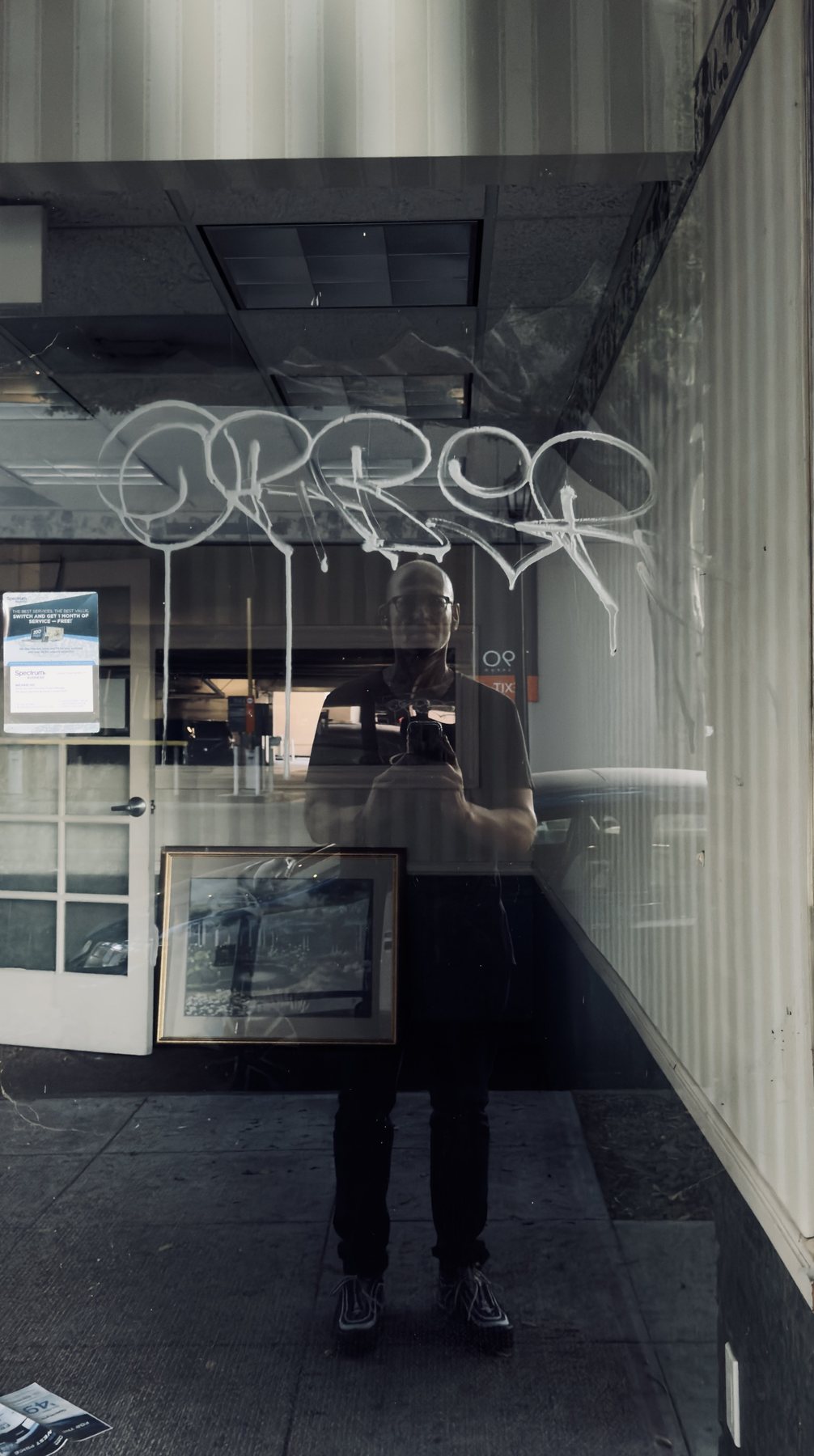 No End In Sight 080: The artist with a camera reflected in a window with white spray-painted tag, a framed picture inside and a street scene behind.