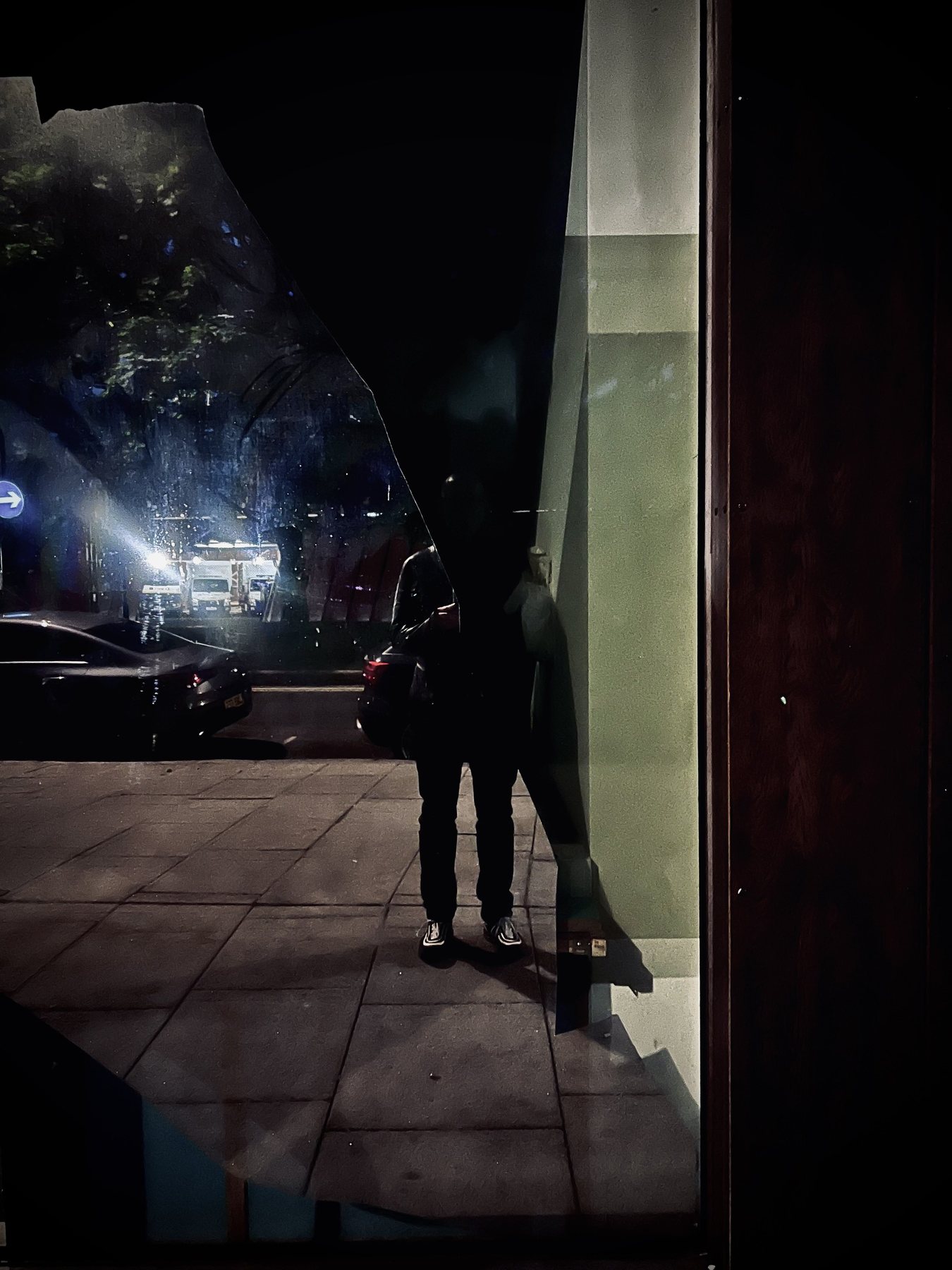 No End In Sight 010: The artist's dark figure in a night window with a broken edge, a pale green wall, a car, blue lights and tree shapes.