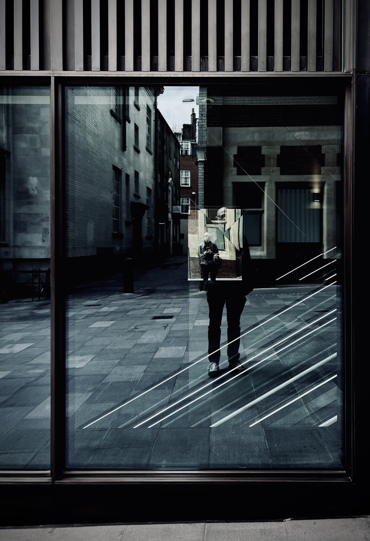 No End In Sight 009: The artist reflected holding a small mirror within the window glass, with white diagonal lines crossing a paved courtyard.
