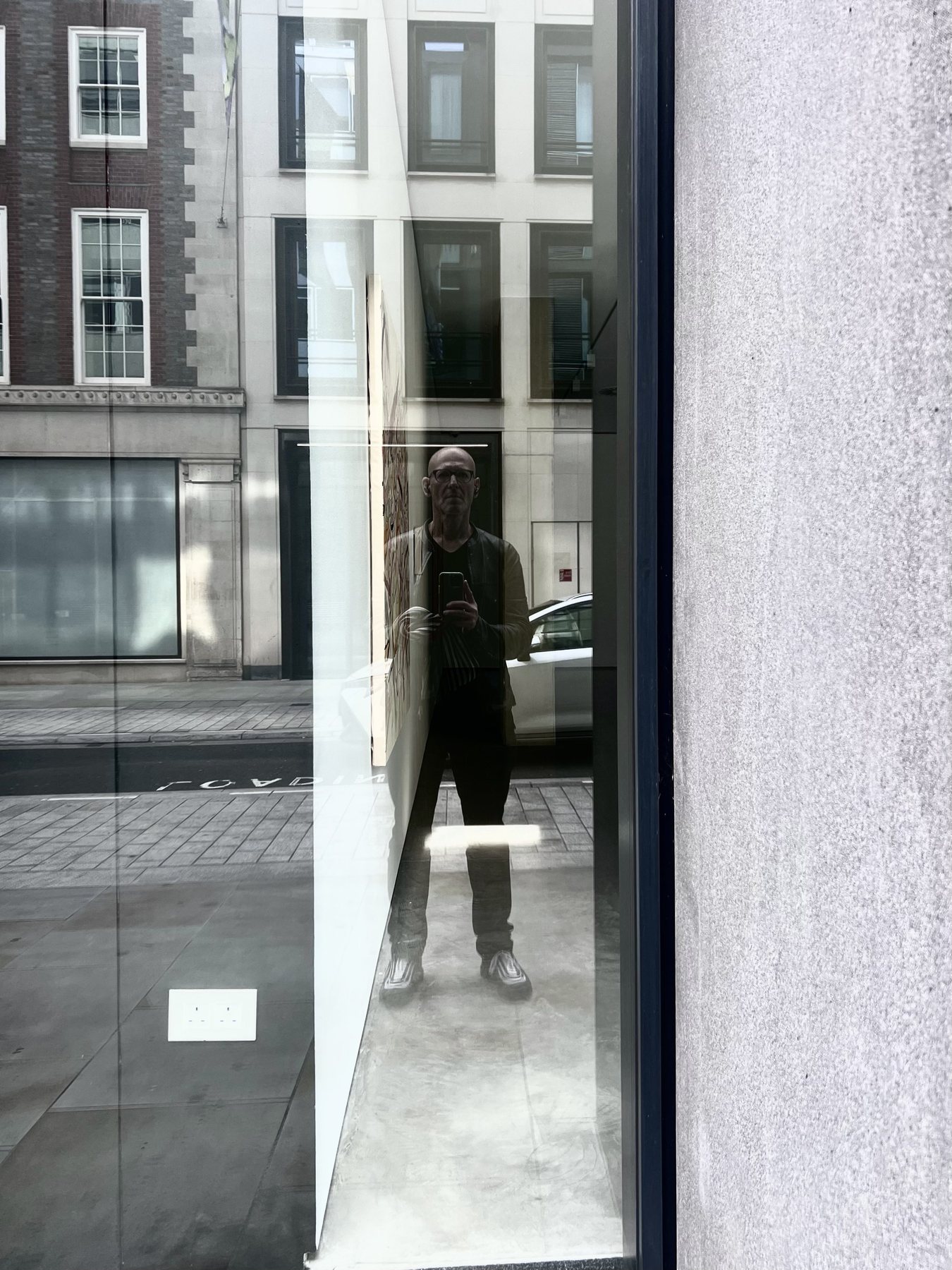 No End In Sight 028: The artist, in glasses holding a phone, reflected full length in a tall window beside a grey stone wall, with terraced buildings behind.
