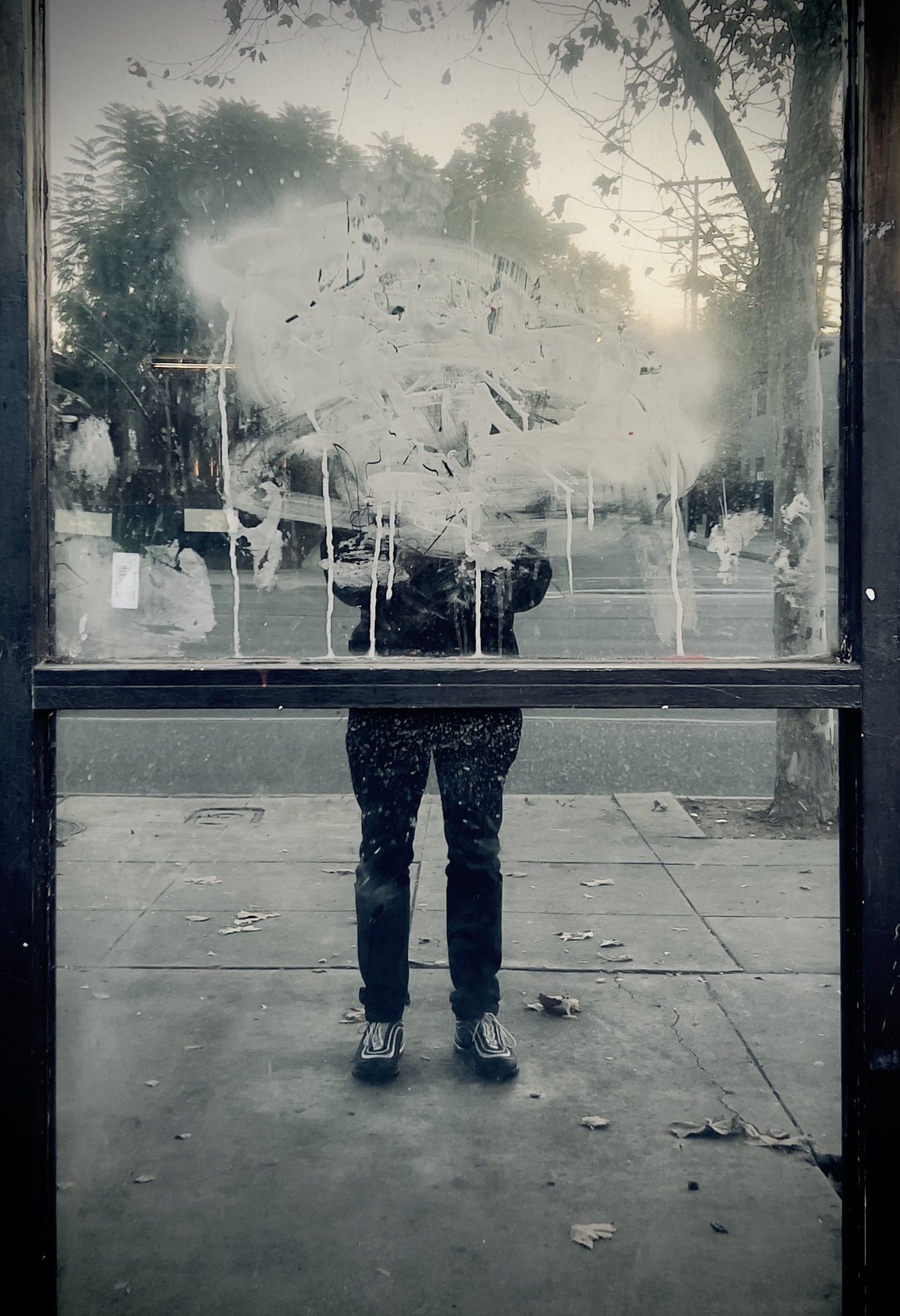 No End In Sight 061: A figure's legs and trainers beneath a smeared white patch on a bus-shelter window, with trees and leaves on the pavement.