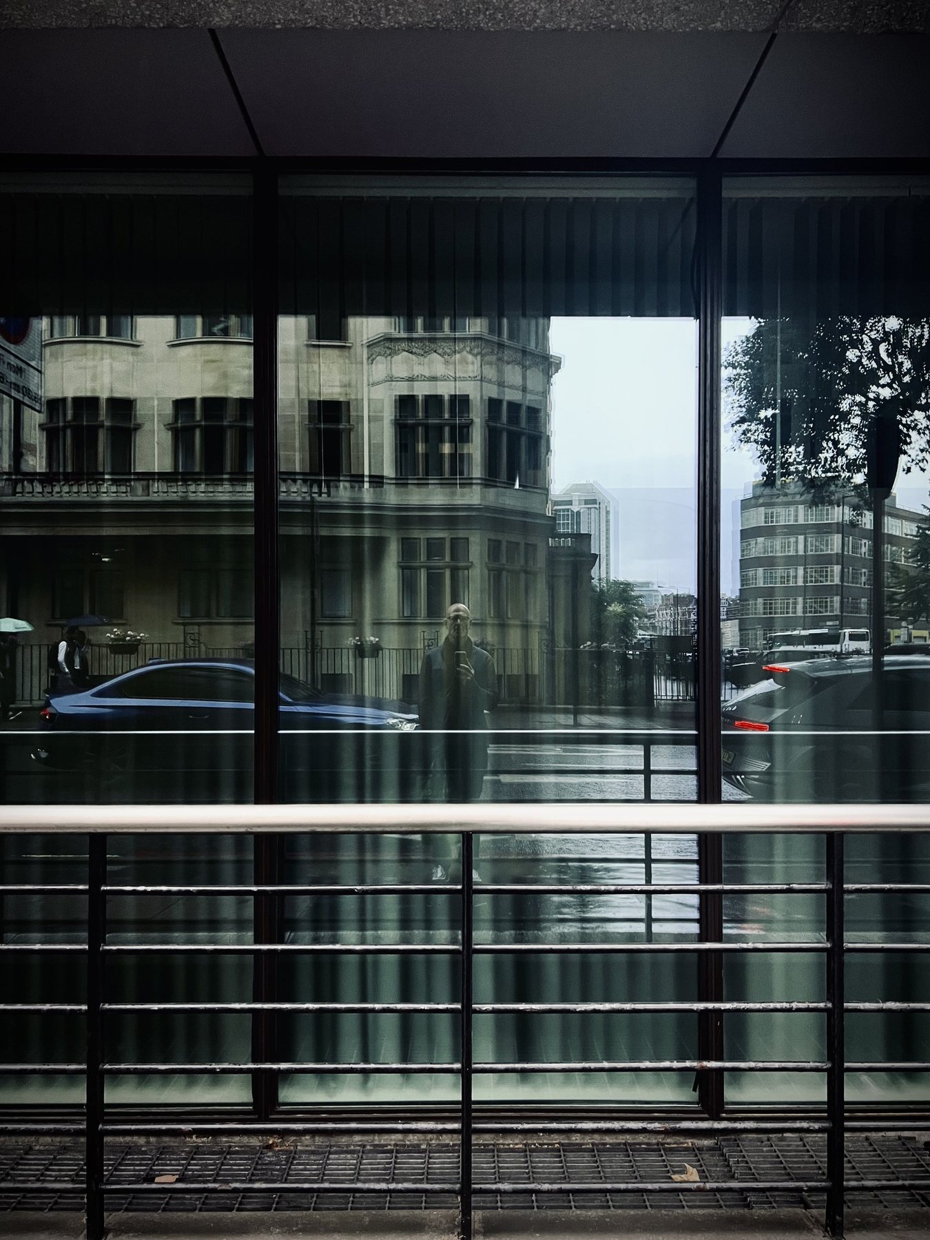 No End In Sight 003: The artist reflected in a rainy-day window with a blue car, a classical building, wet street and a railing in front.
